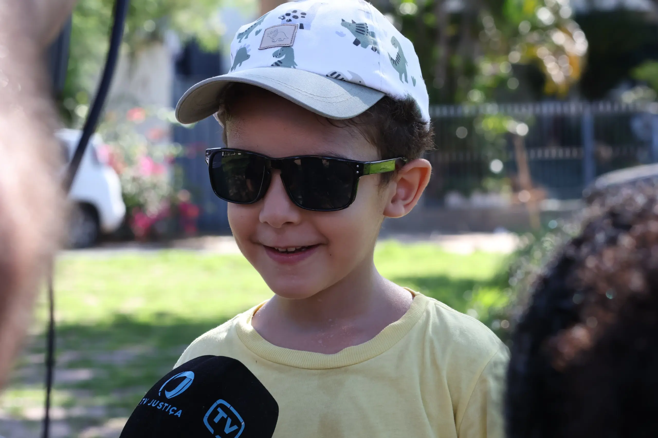 A imagem mostra Raul Benício, 5 anos. Ele é uma criança de pele branca, usa óculos escuros e um boné branco com estampas de dinossauro verde. A sua frente há o microfone da TV Justiça, a qual o jovem concede uma entrevista. Raul está em uma praça com gramados verdes e a luz do sol aparece ao fundo. 