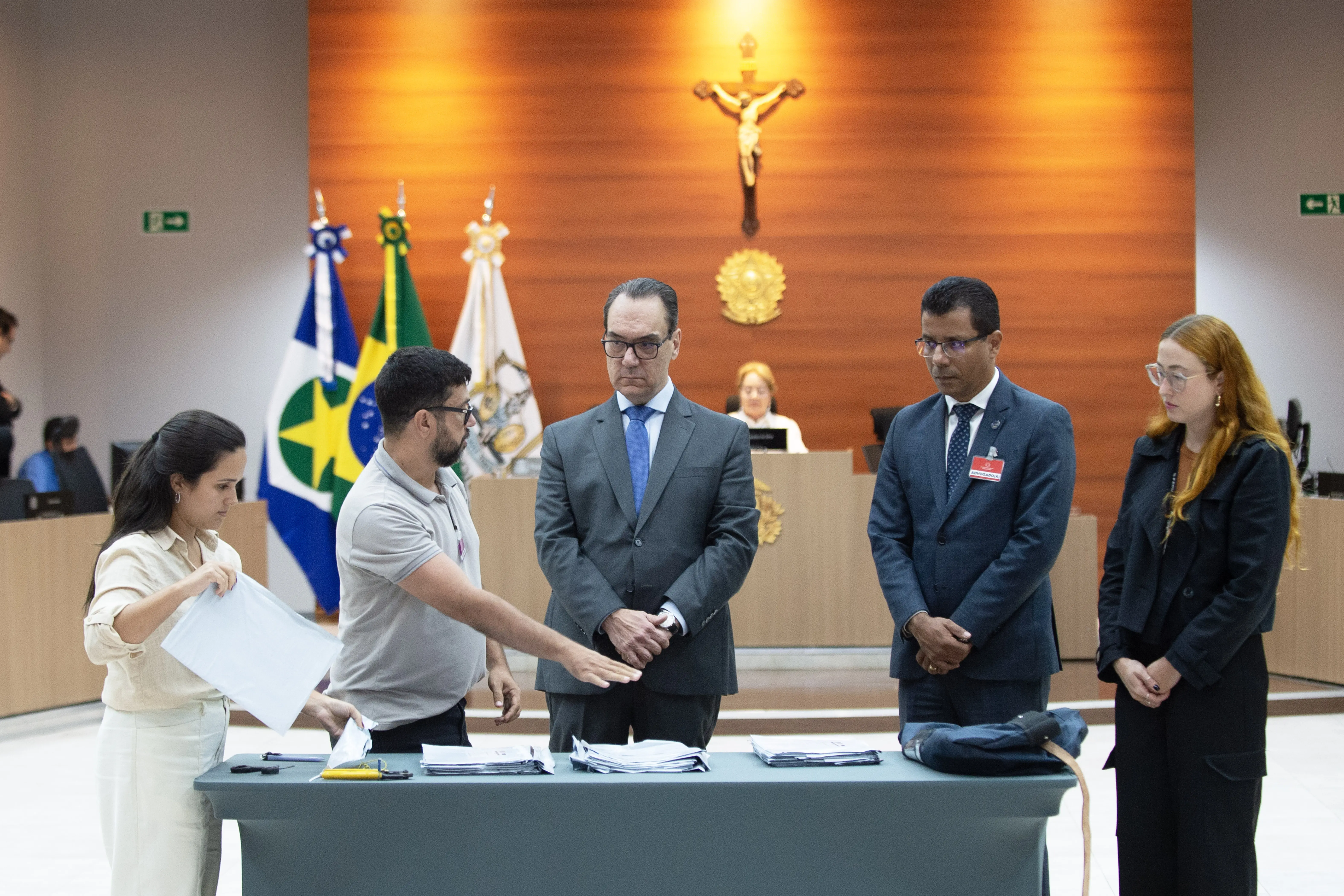 Imagem mostra Plenário 1. Ao centro, uma mesa com malotes. Em volta da mesa, cinco pessoas. Ao fundo, a desembargadora Clarice Claudino preside a sessão de identificação das provas do concurso da magistratura