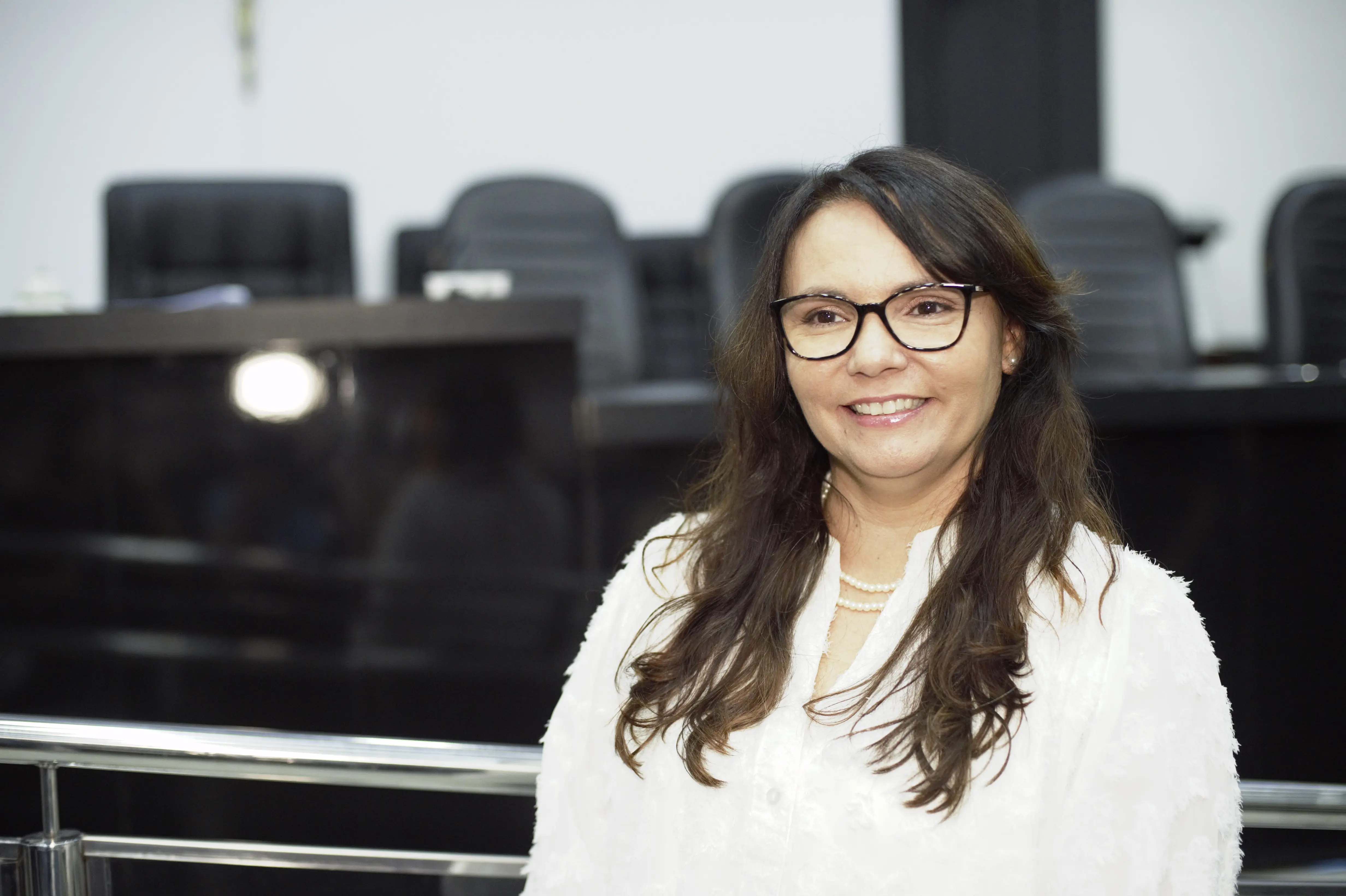 A imagem mostra a presidente da Ampara. Ela é uma mulher de pele clara, cabelos longos, lisos e escuros, olhos escuros e usa óculos de grau, com armação preta. Ela está vestindo uma blusa branca e usa um colar curto de perólas. Está sorrindo