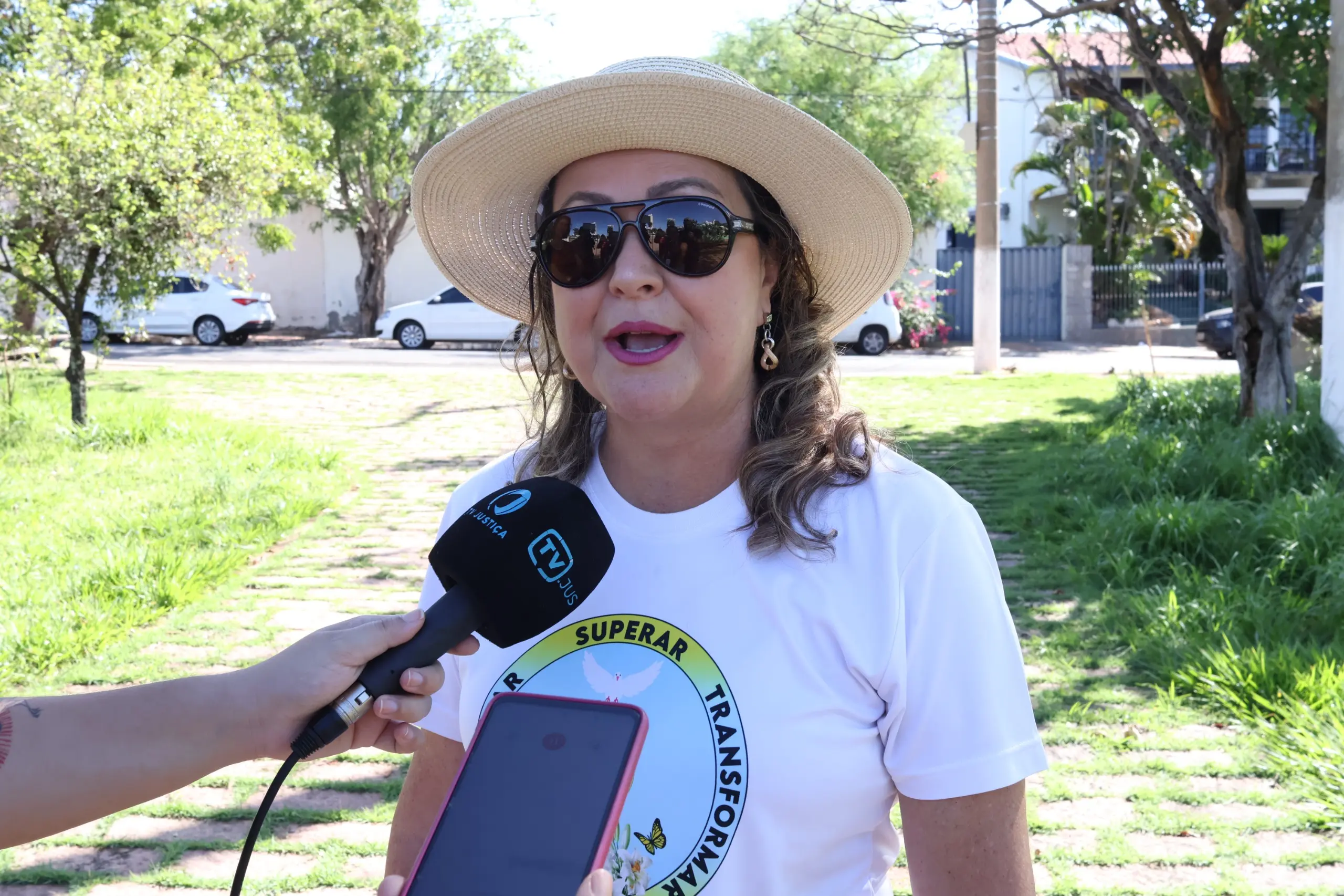 Em destaque está uma Carla Moraes, uma mulher branca de cabelos ondulados até o ombro. Ela usa um chapéu de palha claro, óculos escuros e uma camiseta branca. Carla está em uma praça com gramados verdes e o dia está ensolarado. 