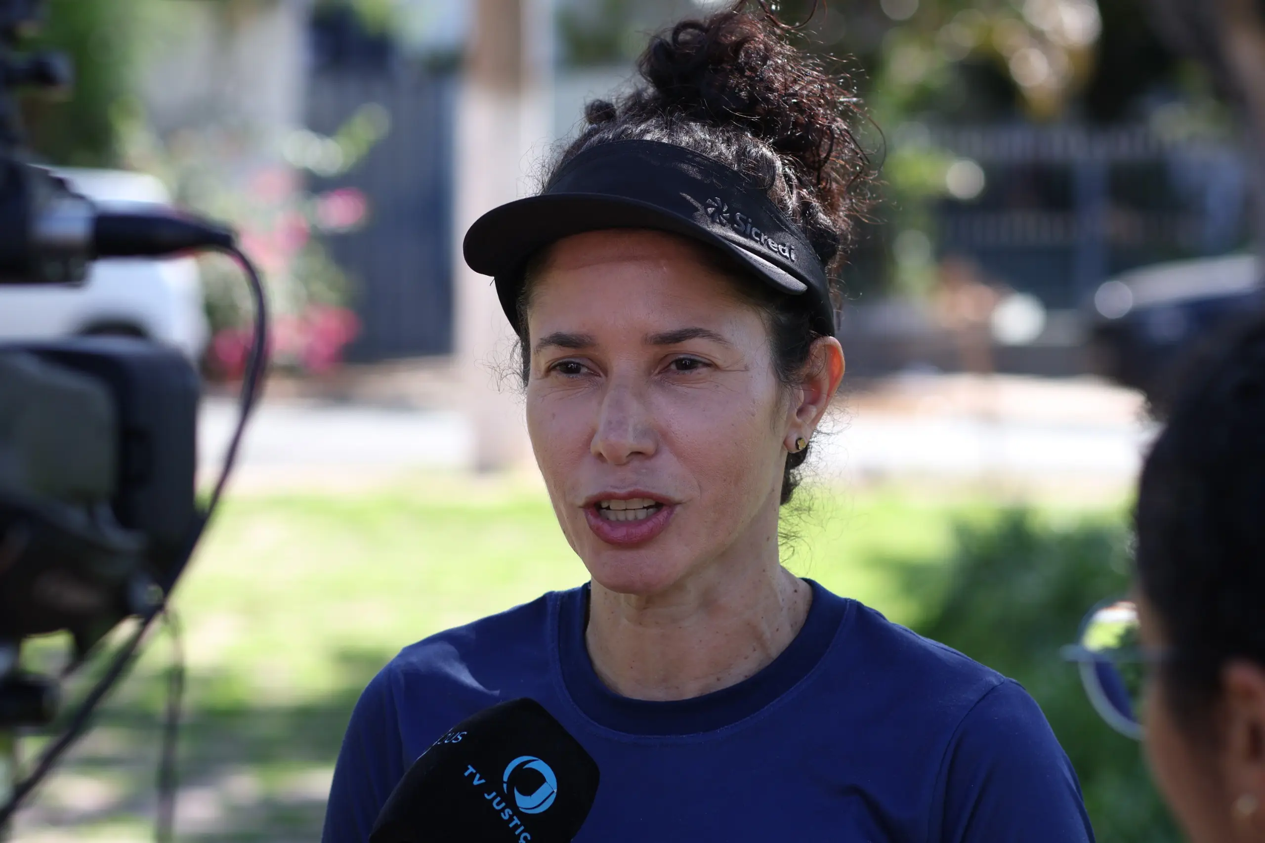 No centro da imagem está Rosiane Carnaíba, uma mulher branca, cabelos cacheados prestos. Ela usa um boné de aba preto e os cabelos estão preso em um coque no alto da cabeça. Rosiane usa uma camiseta azul escura e a sua frente há um microfone da TV Justiça, a qual ela concede uma entrevista. 