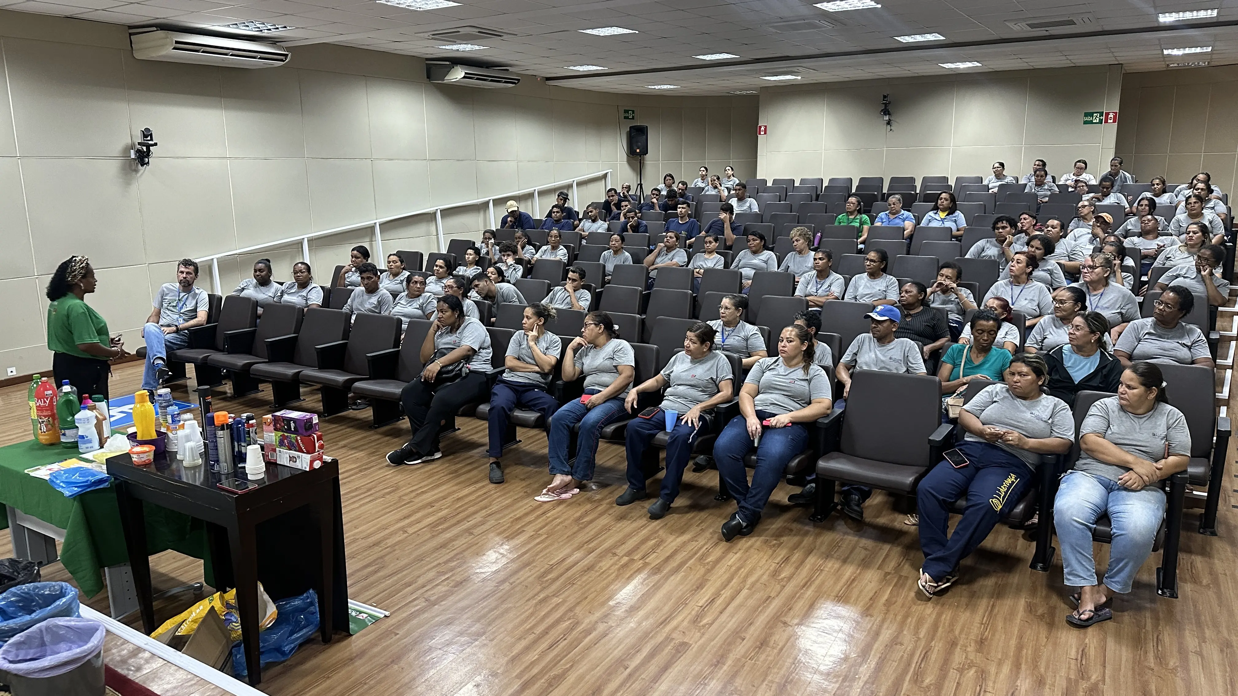 Foto em plano aberto que mostra o auditório do TJMT lotado de funcionários terceirizados assistindo a palestra da educadora ambiental Cidinha Nascimento, que está a frente, falando em pé. Cidinha é uma mulher negra usando camiseta verde da Asmats e faixa no cabelo. Ao lado dela, há uma mesa repleta 