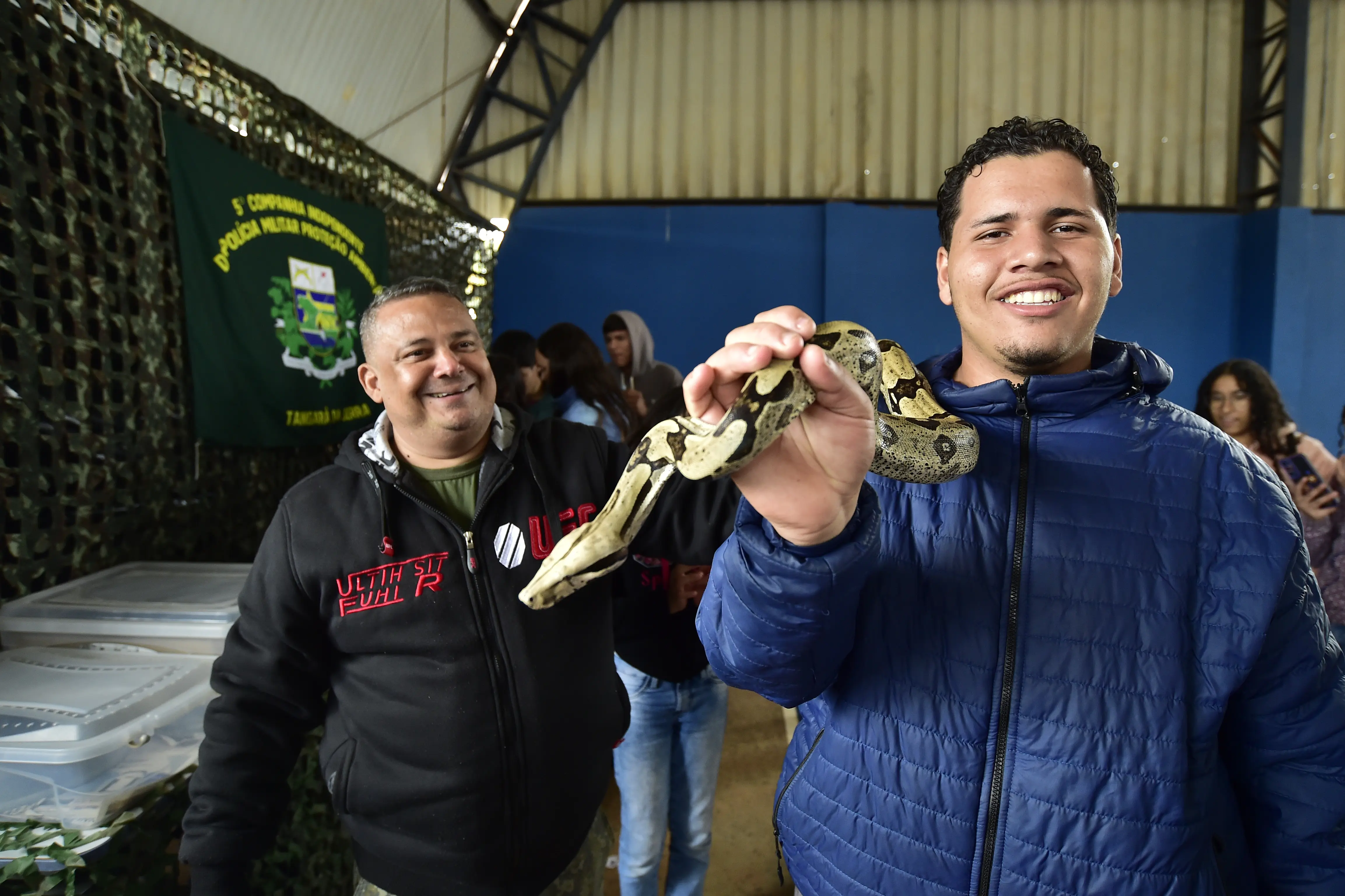 Adolescente segura uma cobra e a mostra, sorrindo, para a foto, em uma quadra esportiva. Ao lado dele, está o policial ambiental R. Rodrigues, sorrindo.