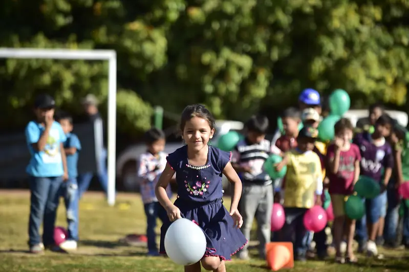 Em um campo de futebol, várias crianças participam de uma gincana. Em primeiro plano, uma menina corre segurando um balão.