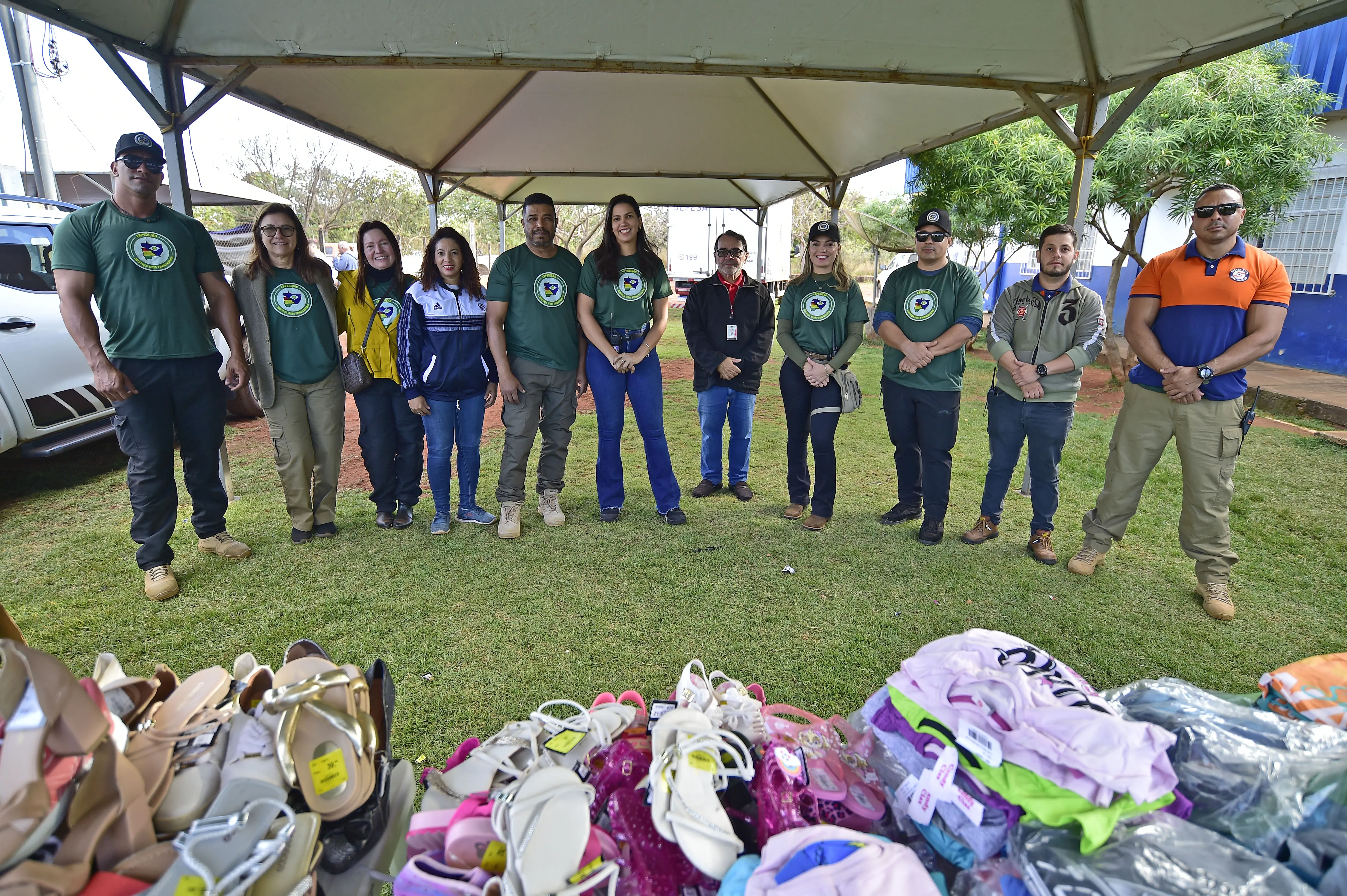 Embaixo de uma tenda, no pátio de uma escola, voluntários da Expedição Justiça Sem Fronteiras posam para a foto em frente a uma mesa cheia de roupas e calçados para doação.