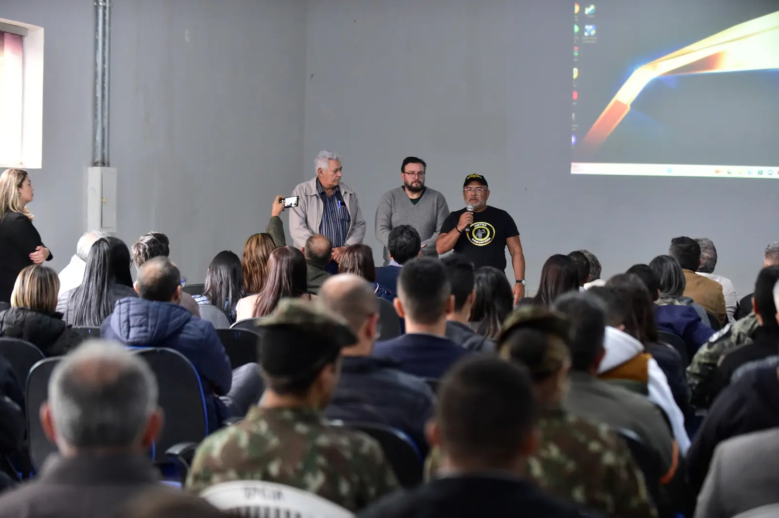 Auditório com público atento. Três homens estão em pé à frente, um deles fala ao microfone usando camiseta da Justiça Comunitária. Telão exibe tela inicial de computador. Pessoas sentadas assistem à fala.