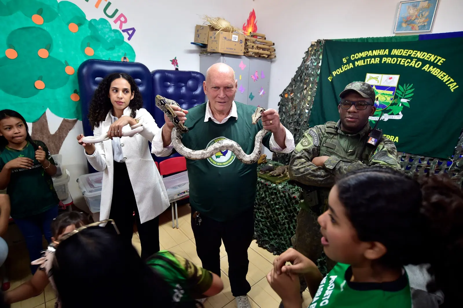 Em uma sala de aula, o prefeito de Vila Bela da Santíssima Trindade, André Bringsken, e uma médica seguram uma enorme cobra branca e a exibem para a foto, ao lado de um policial ambiental. Há crianças em volta.