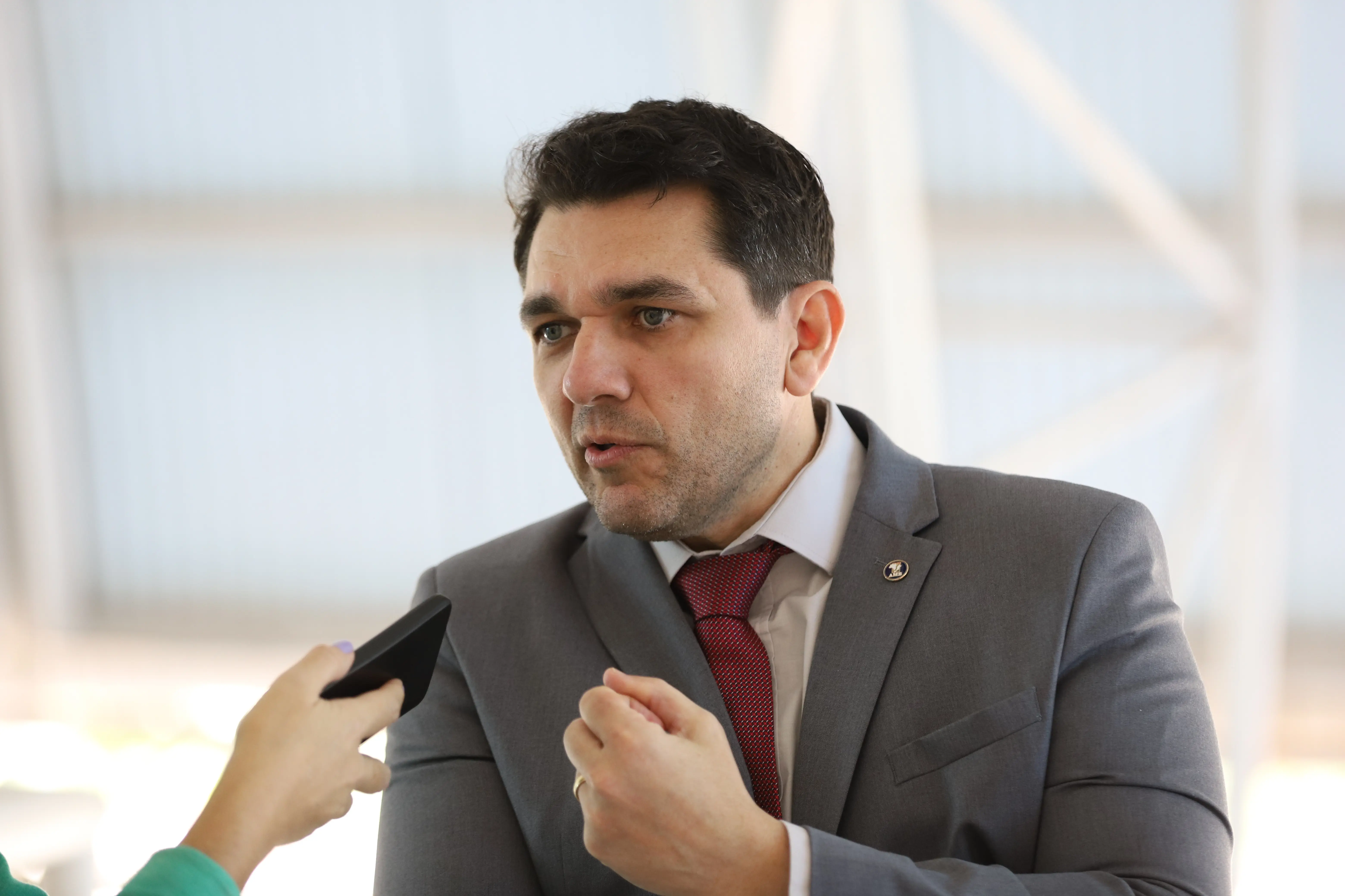 Um homem de terno cinza e gravata vermelha, Decildo, está sendo entrevistado. Ele gesticula e olha para o lado, enquanto uma mão segura um microfone. O fundo é claro e desfocado.