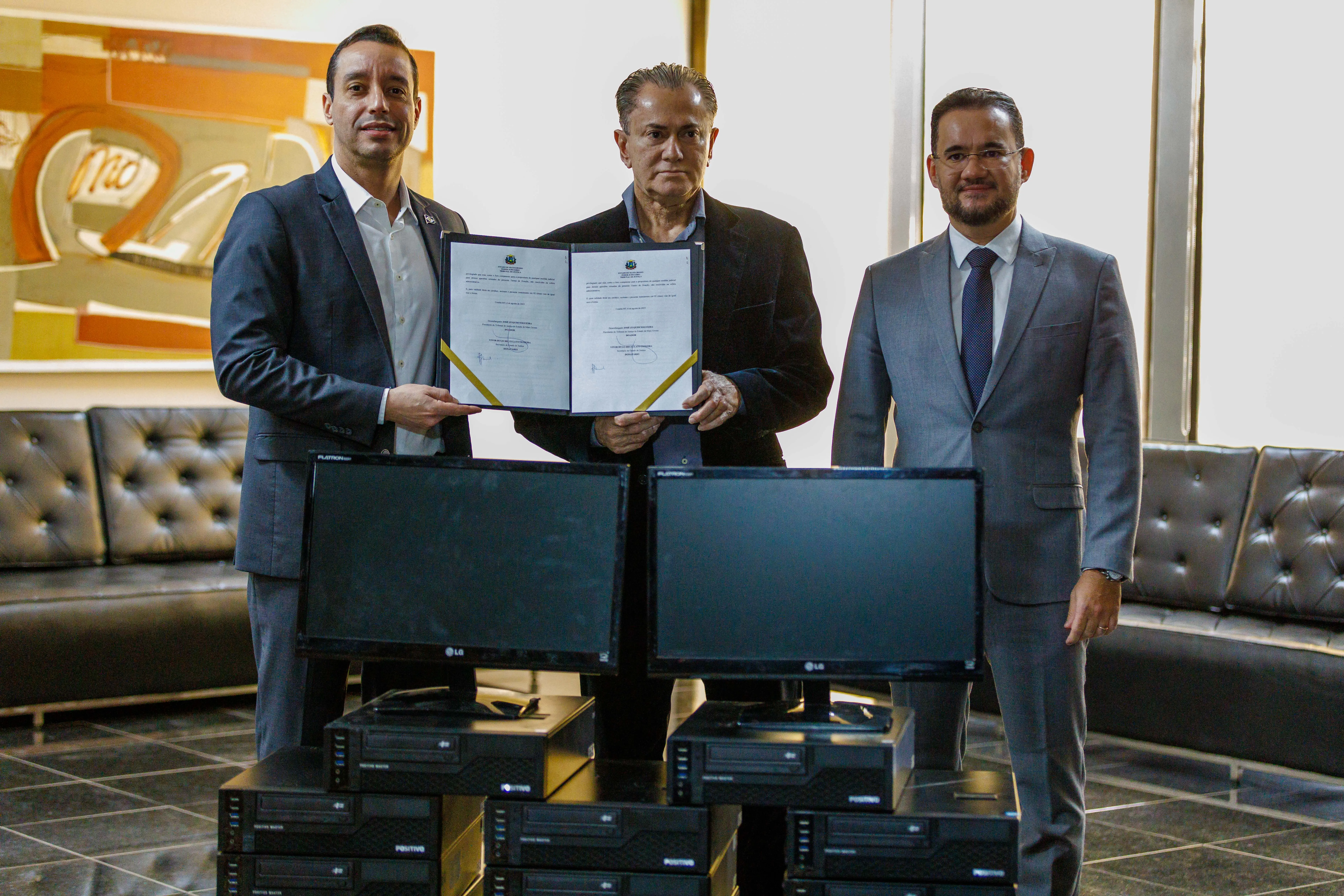 Três homens de terno posam em uma sala. O homem do centro segura um documento, enquanto os outros dois seguram cada um dos lados do documento, com computadores à frente.