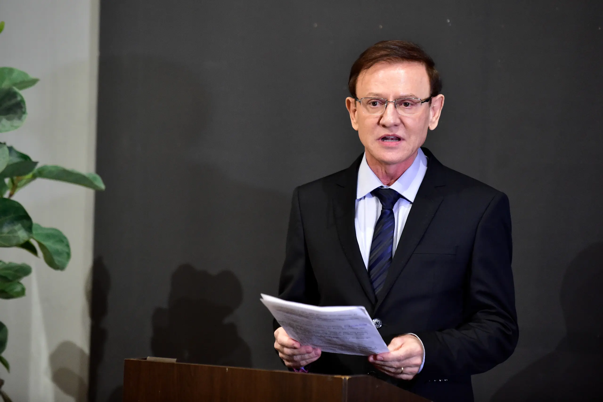 Foto colorida e horizontal. Juiz Wanderlei dos Reis usa terno e gravata preto, além de camisa branca. Ele tem cabelos castanhos e usa óculos. Está atrás de um púlpito, segura um papel às mãos e está falando