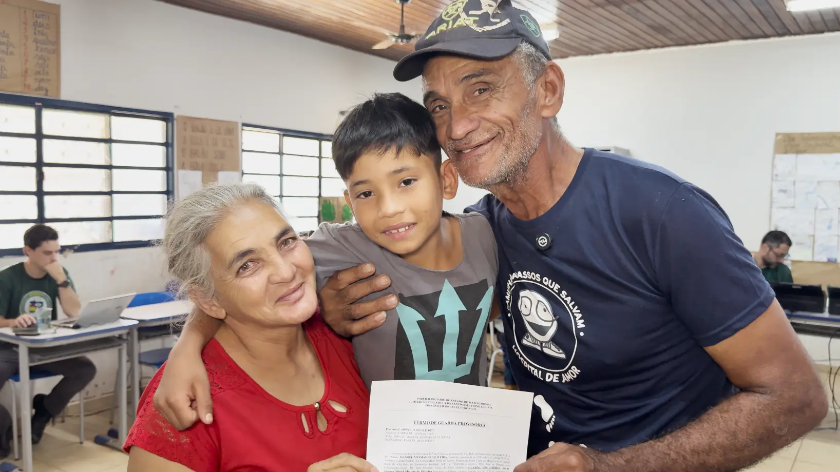 Foto que mostra o menino Gabriel sendo abraçado pela avó e pelo avô maternos. Eles sorriem para a foto. Os avós mostram o documento da guarda do menino.