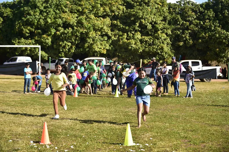 Em um campo de futebol, várias crianças participam de uma gincana. Em primeiro plano, duas meninas correm segurando um balão, cada, em direção a cones.