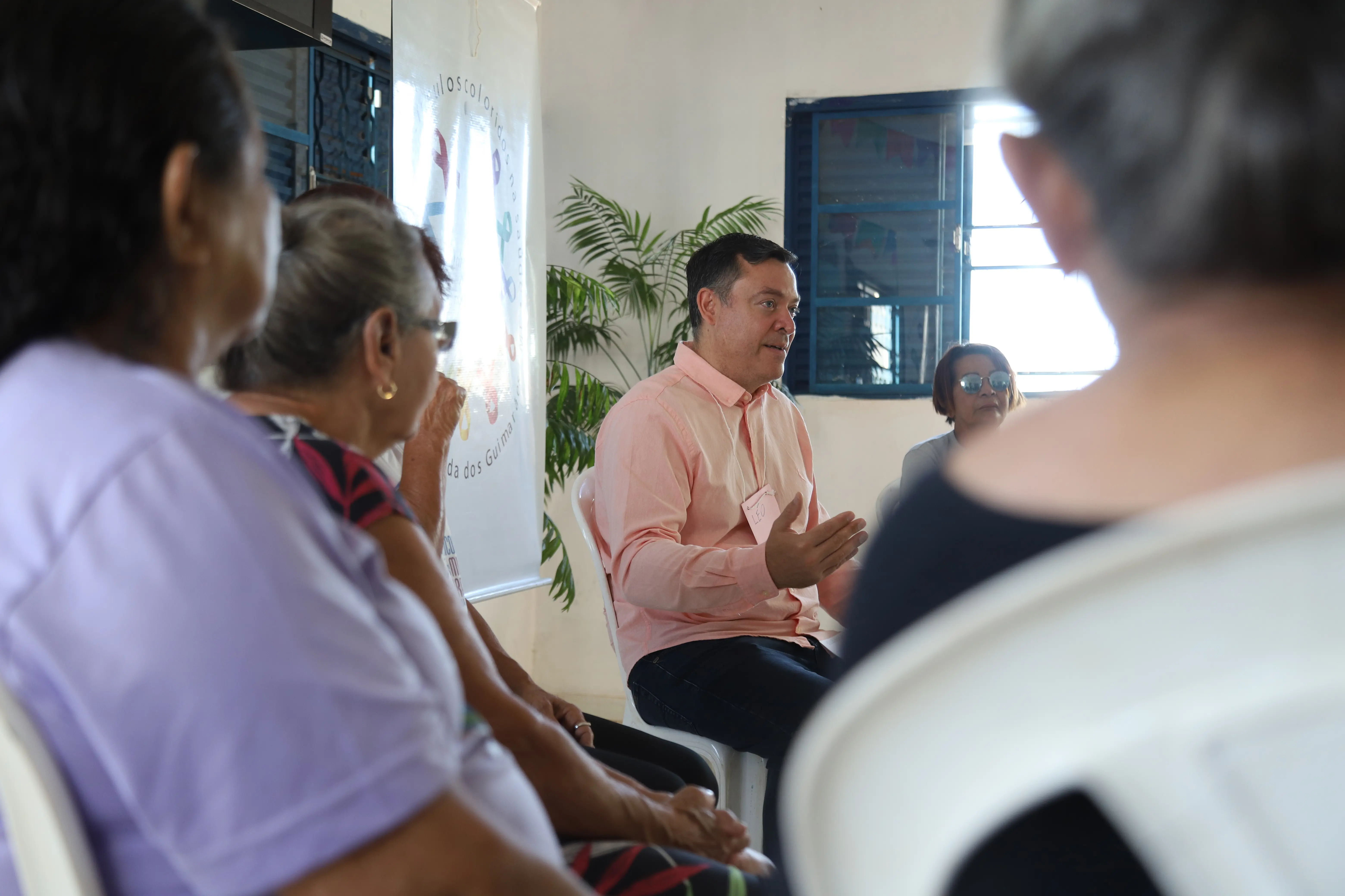 Facilitador, juiz Leonísio Salles, conduz roda de conversa com pessoas idosas em círculo, promovendo escuta ativa e diálogo. Ao fundo, banner do projeto “Círculos Coloridos na Saúde” e janela com iluminação natural.