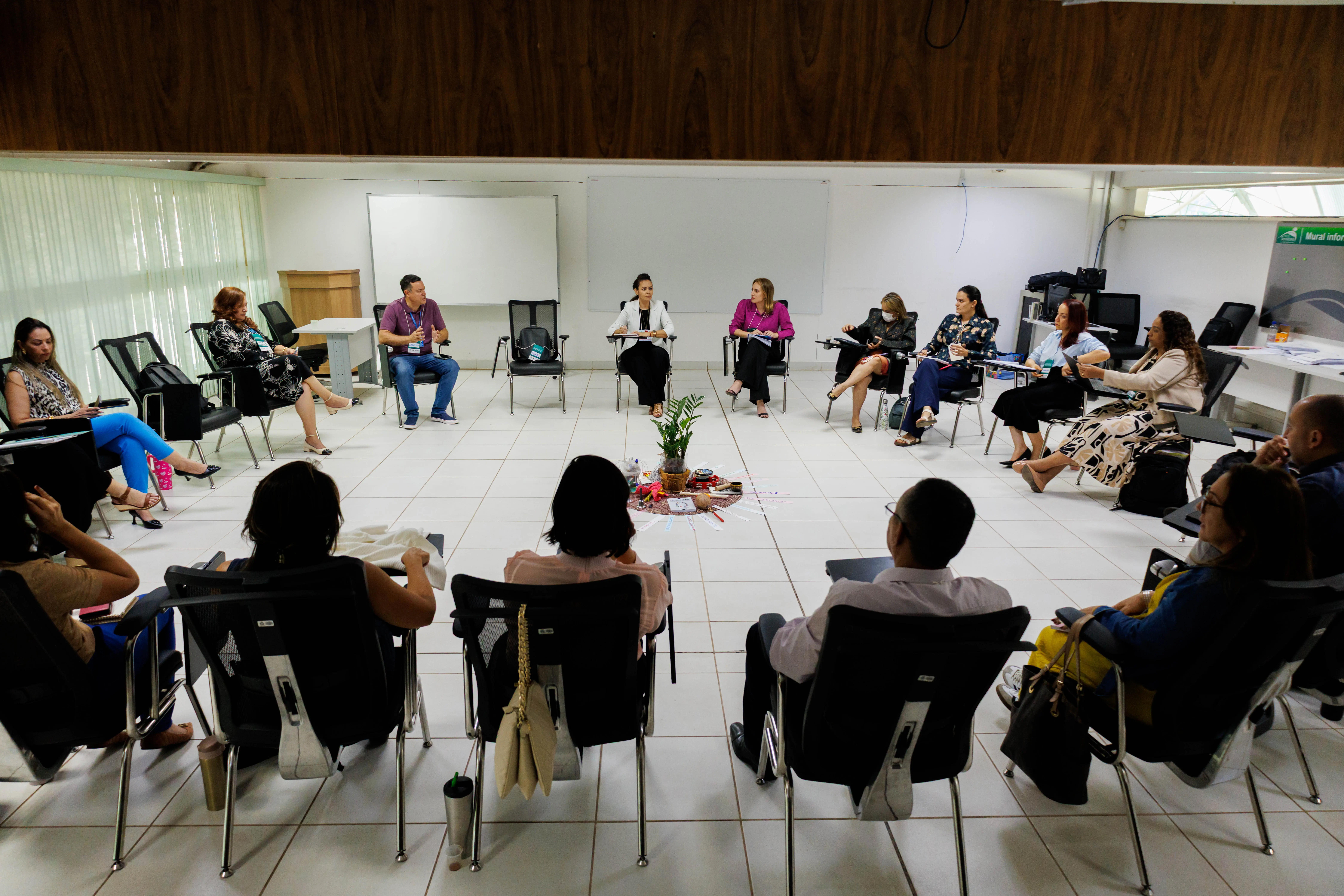 A imagem retrata uma reunião em sala iluminada, com pessoas sentadas em círculo. No centro há objetos e uma planta. Algumas anotam ou falam; há janelas grandes e um quadro branco ao fundo.