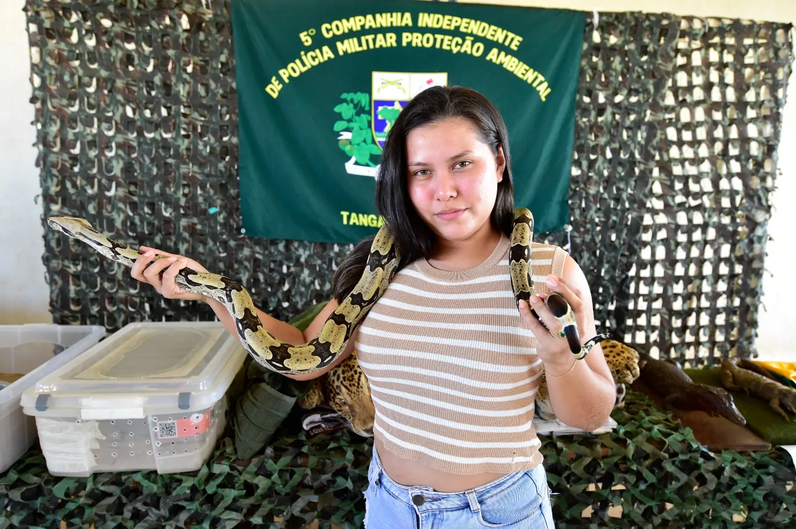 Menina adolescente segura uma cobra e a mostra, sorrindo, para a foto, em uma quadra esportiva. Atrás dela, está o estande da Polícia Ambiental.