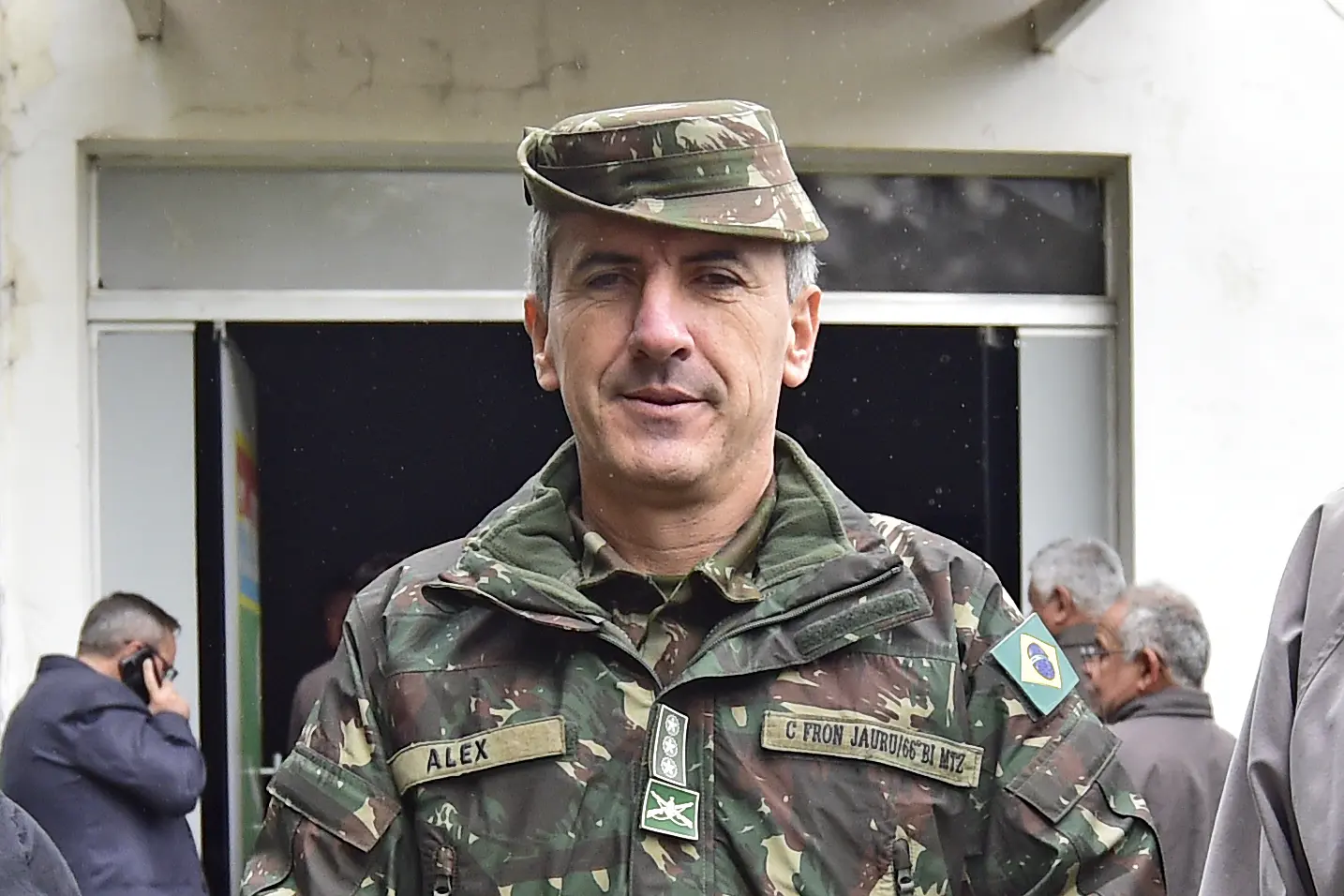 Militar fardado, com expressão séria e uniforme camuflado do Exército Brasileiro, posa para foto em frente a prédio branco. Ao fundo, homens conversam e um deles fala ao celular próximo à entrada.