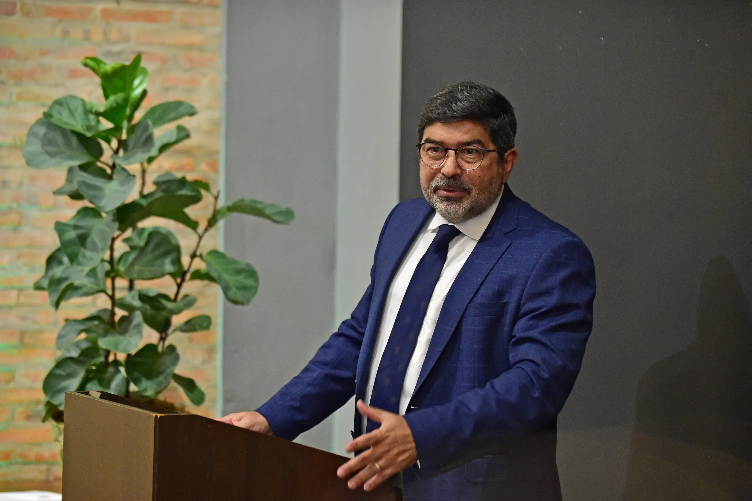 Foto colorida e horizontal. Juiz Antônio Horácio veste paletó e gravatas azul, camisa branca. Ele tem cabelos curtos e pretos e usa óculos. Atrás dele, em segundo plano, está uma planta com folhas grandes