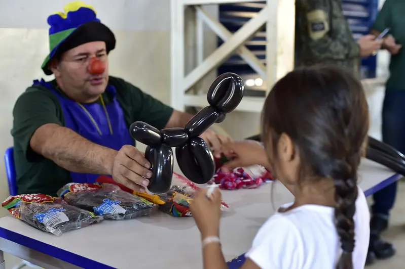 Palhaço Lelé Picole Curimpampam entrega um bichinho de balão preto para uma menina, que está de costas na foto.