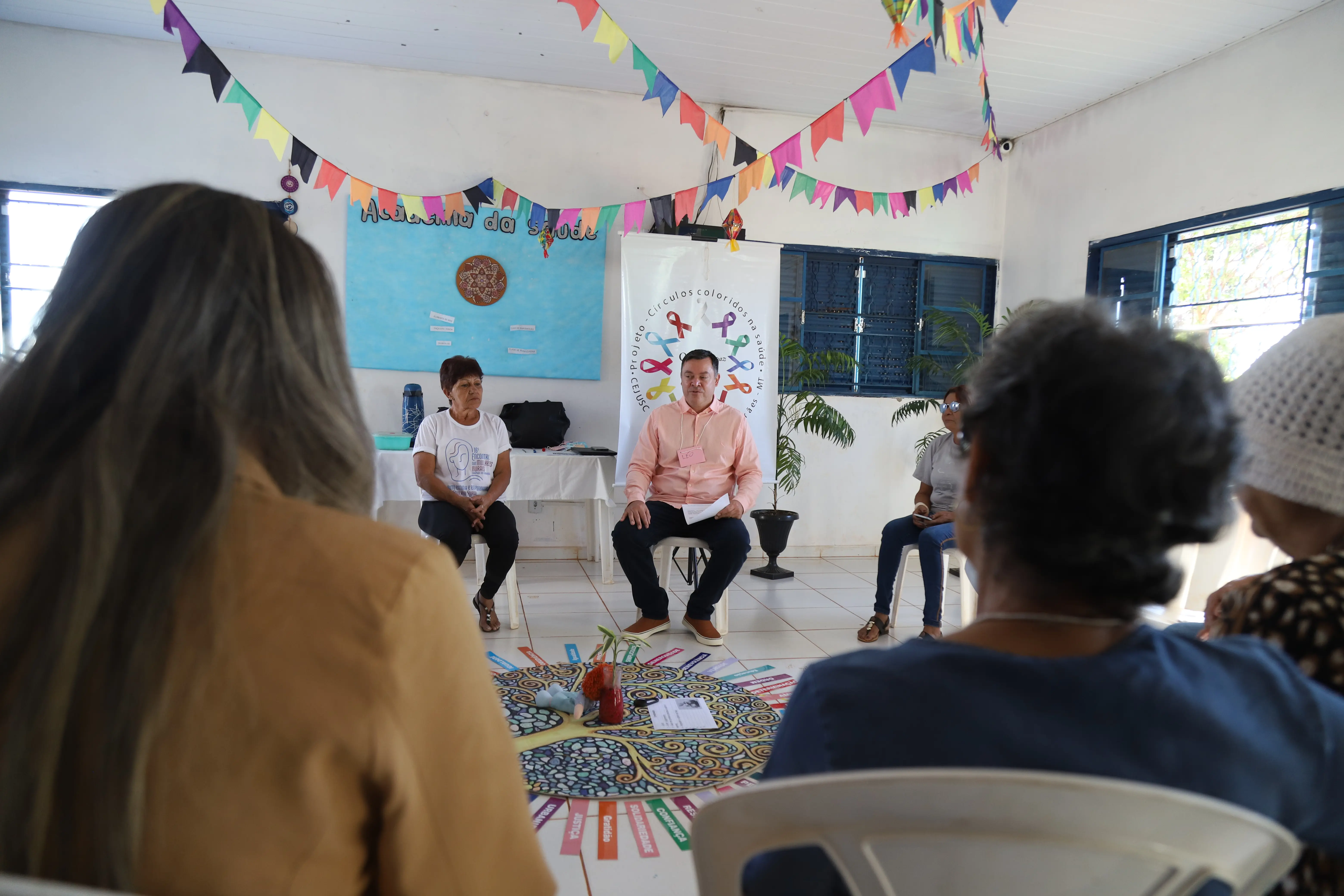 Círculo de Construção de Paz acontece em espaço decorado com bandeirinhas coloridas. Facilitador e grupo dialogam em ambiente leve, com foco em fortalecimento de vínculos e valores comunitários