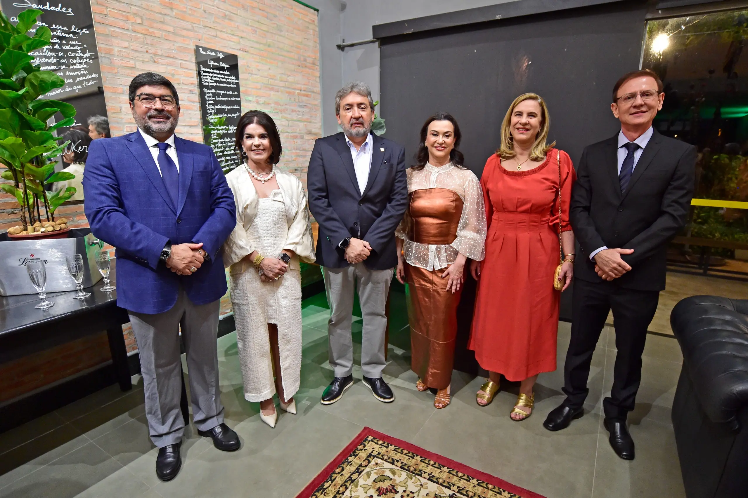 Foto colorida e horizontal. Seis pessoas estão em pé, uma ao lado da outra, posam e sorriem para a foto. Elas usam roupas coloridas