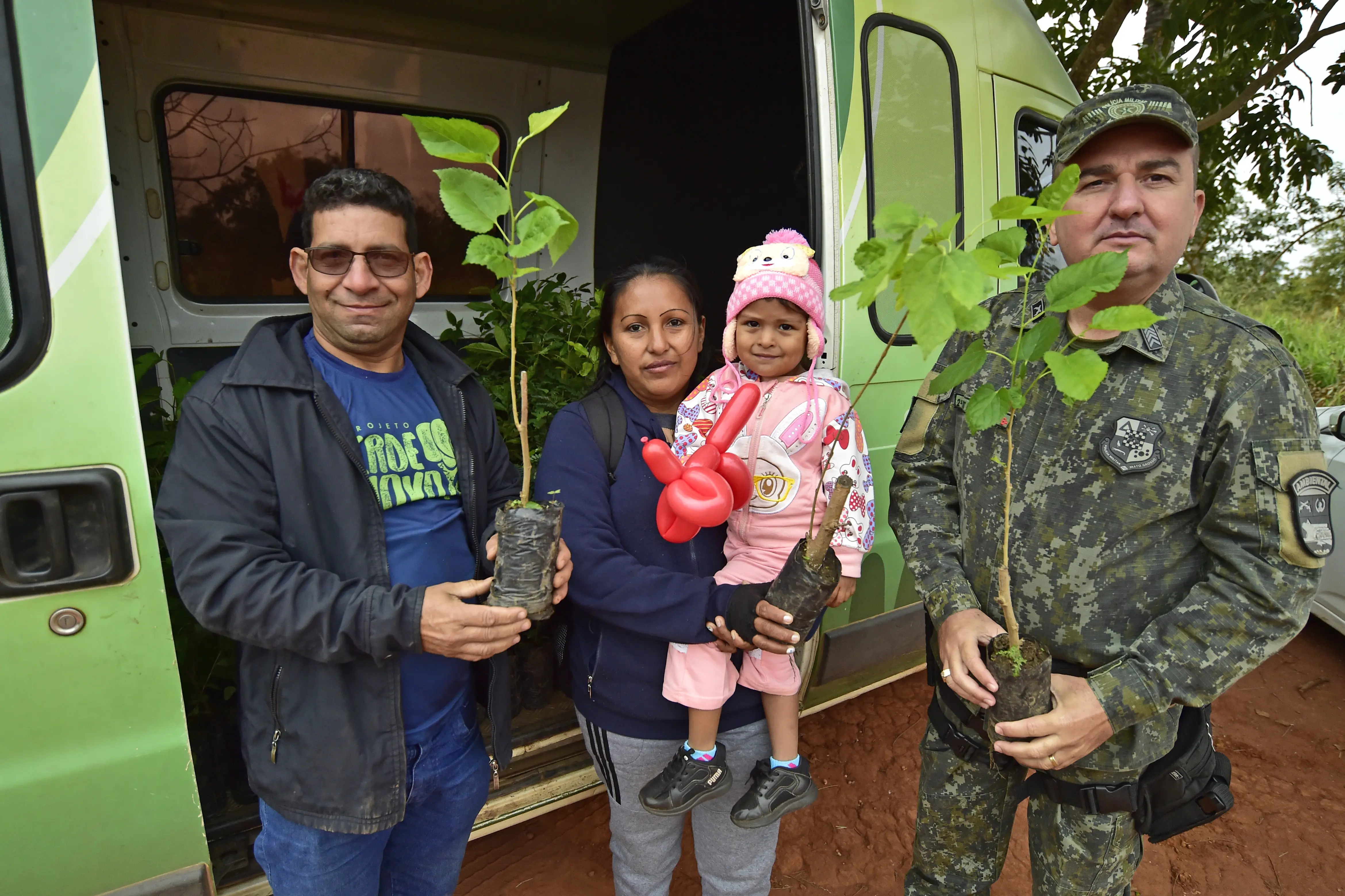 Servidor com a camiseta do programa Verde Novo, mulher com criança no colo e policial ambiental posam para a foto, em frente à van do programa Verde Novo. Eles seguram mudas de plantas;