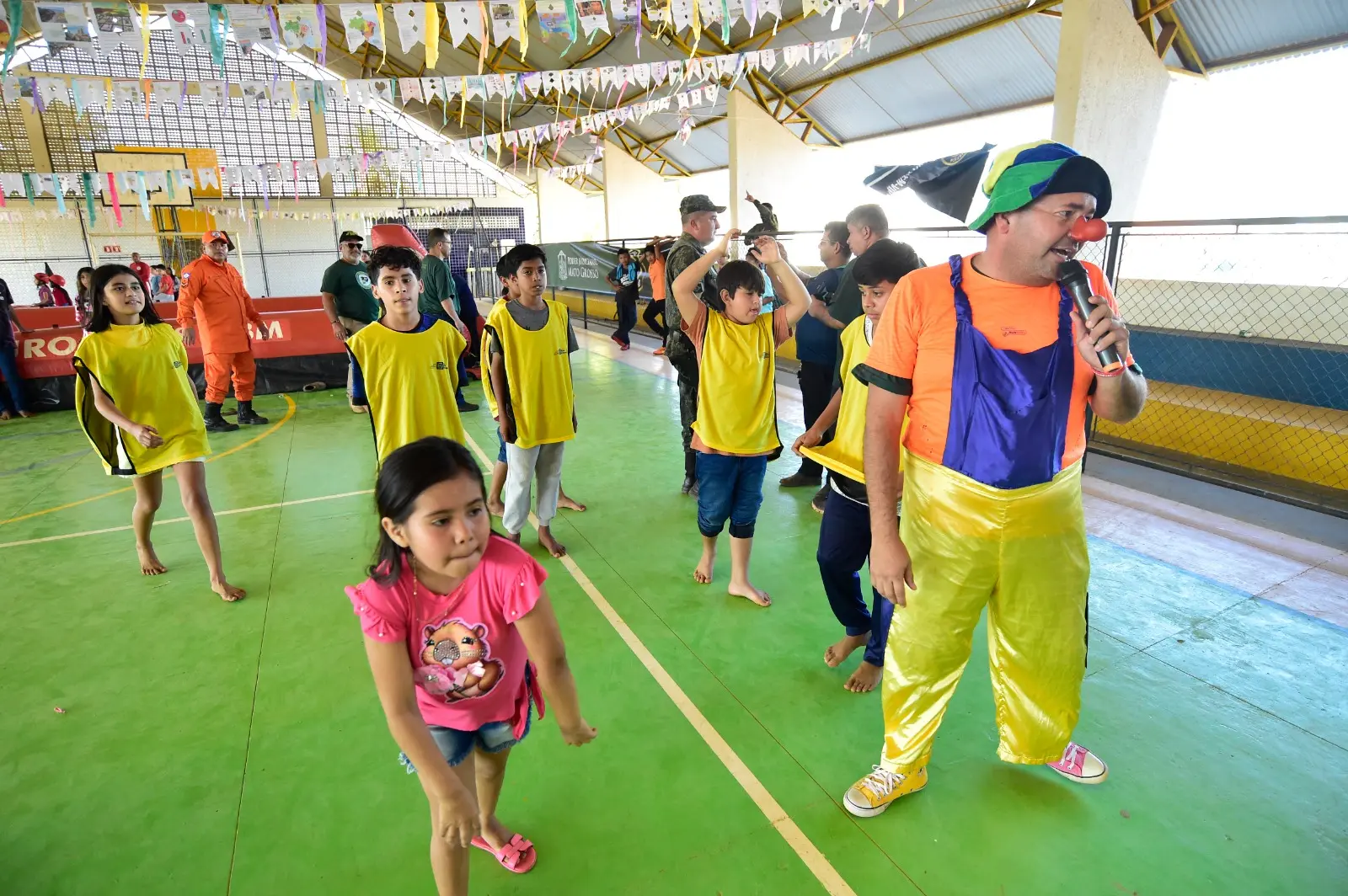 Em uma quadra esportiva, o palhaço Lelé Picolé Curimpampam conduz uma gincana entre crianças.