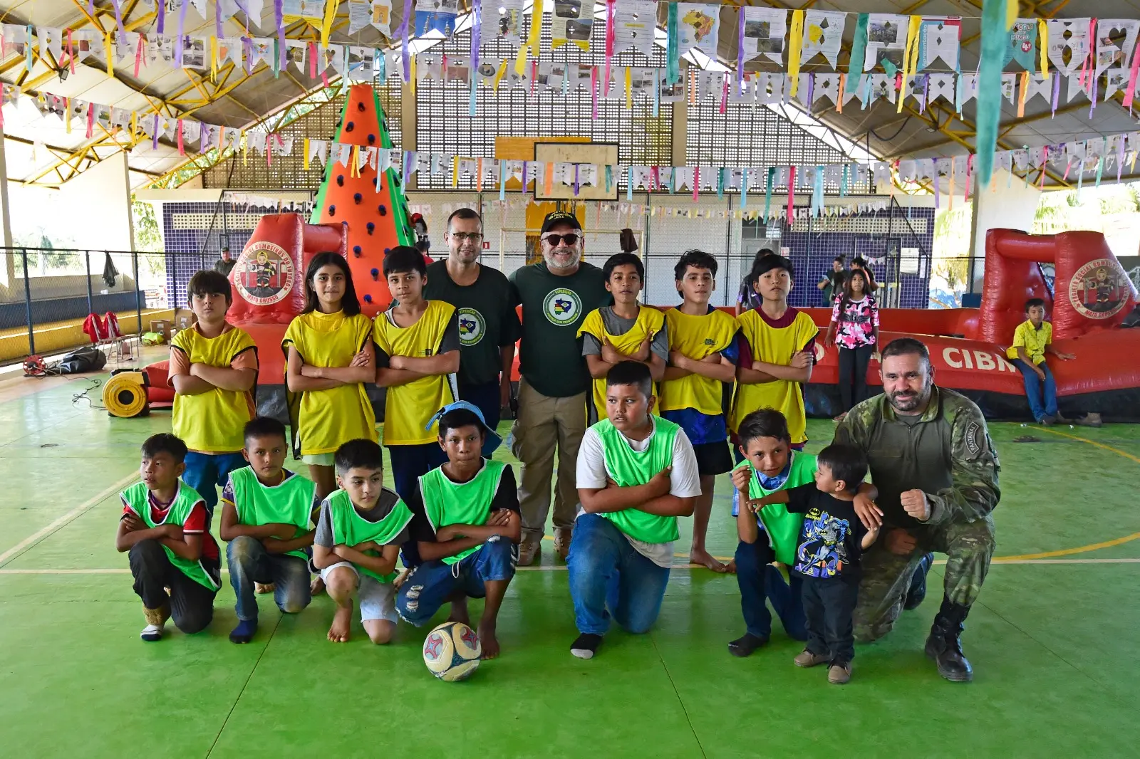 Em uma quadra esportiva, várias crianças de dois times diferentes, com coletes verde e amarelo, posam em posição de pré-jogo, junto com o prefeito de Porto Esperidião, o juiz José Antônio Bezerra Filho e um policial do Gefron