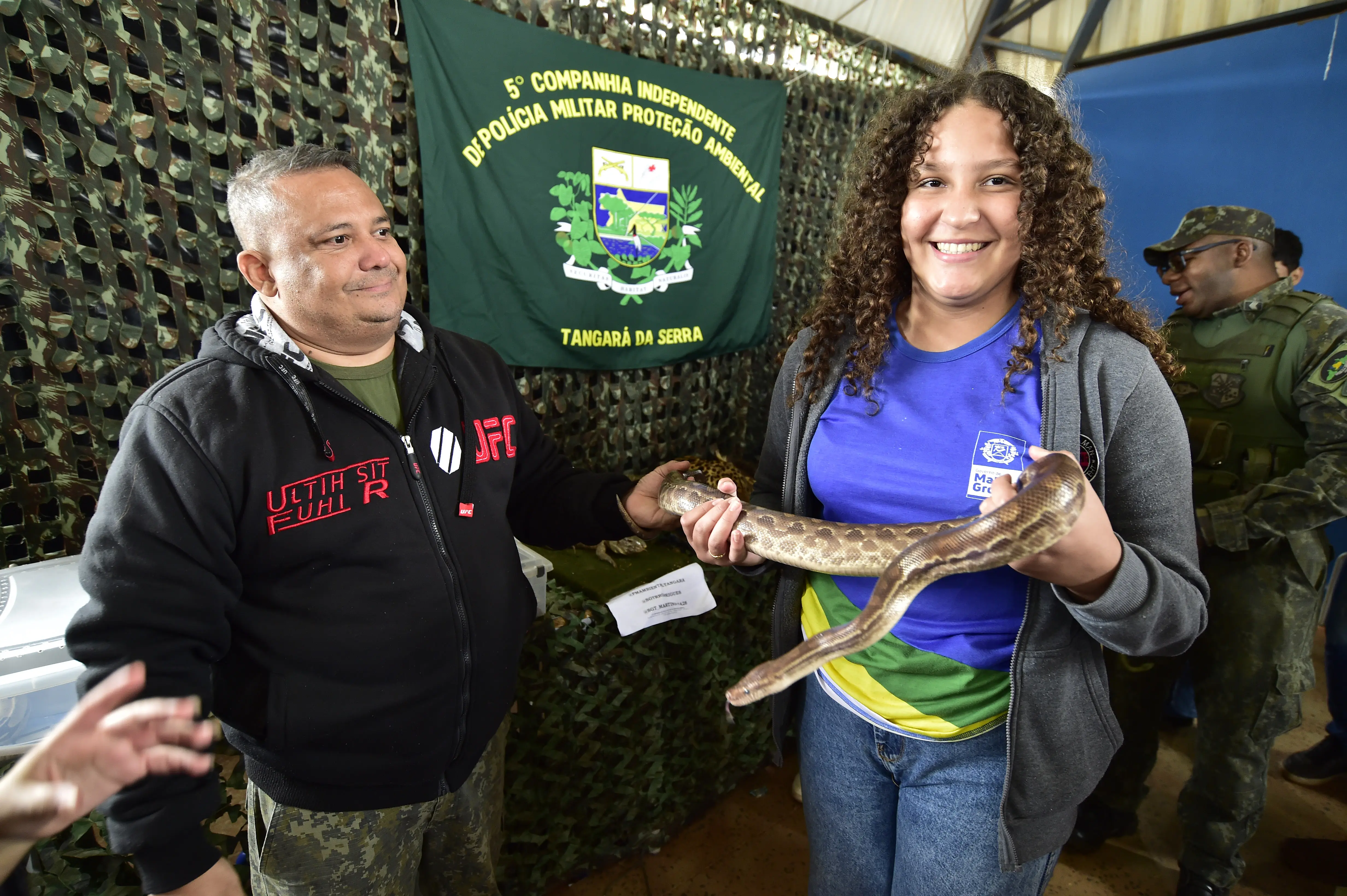 Menina adolescente segura uma cobra e a mostra, sorrindo, para a foto, em uma quadra esportiva. Ao lado dela, está o policial ambiental R. Rodrigues, sorrindo.