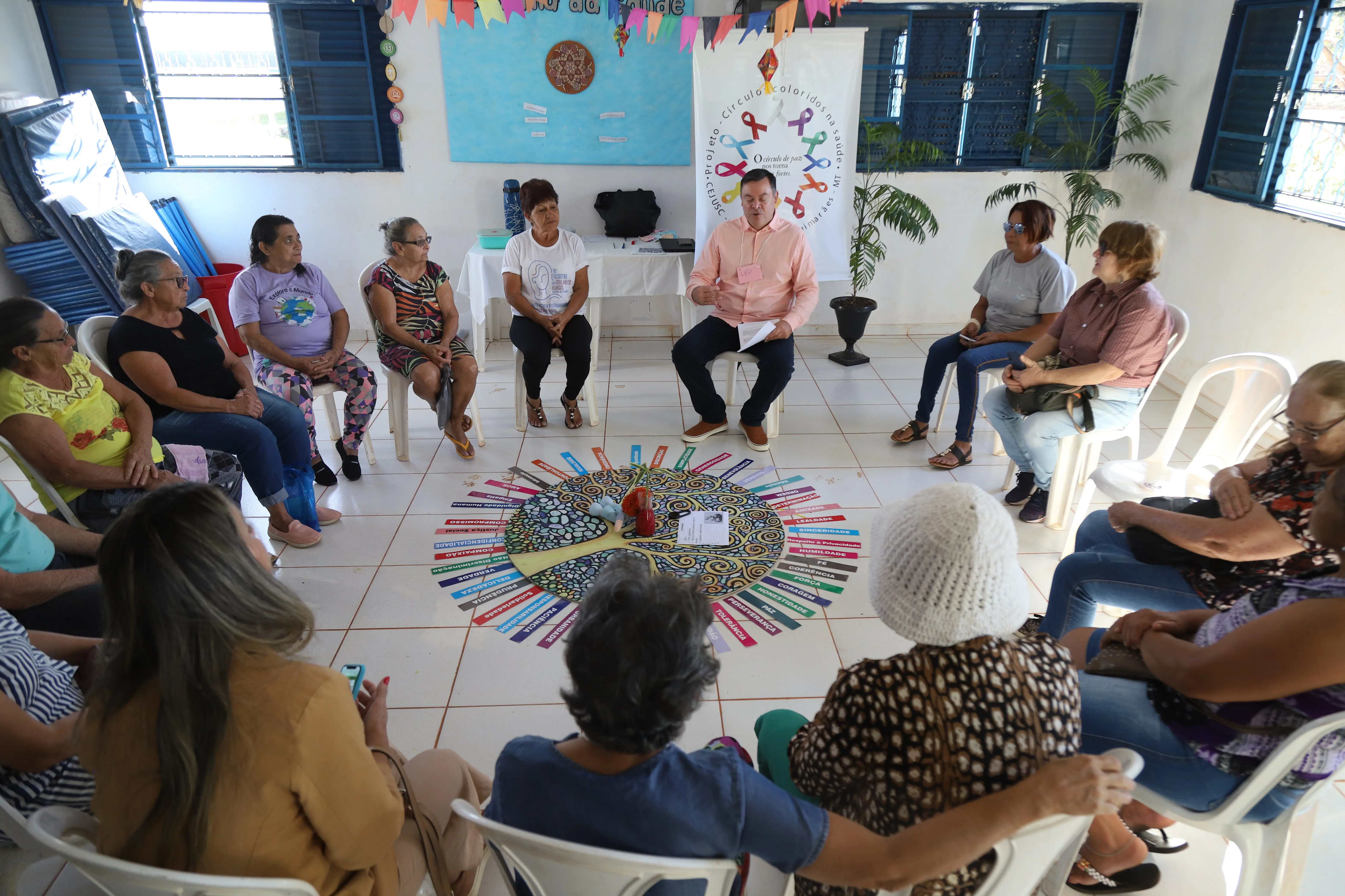 Participantes formam círculo ao redor de mandala com palavras de valores humanos. Ambiente decorado com bandeirolas e cartazes, reforçando clima acolhedor e colaborativo do encontro