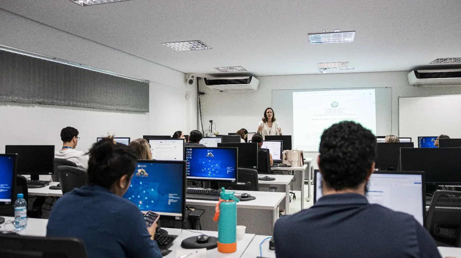 Foto em plano aberto, que mostra a diretora do DAJE, Shusiene Tassinari Machado em pé, à frente da sala, conduzindo a aula enquanto os participantes acompanham a apresentação em computadores no Laboratório da Escola dos Servidores do Poder Judiciário.