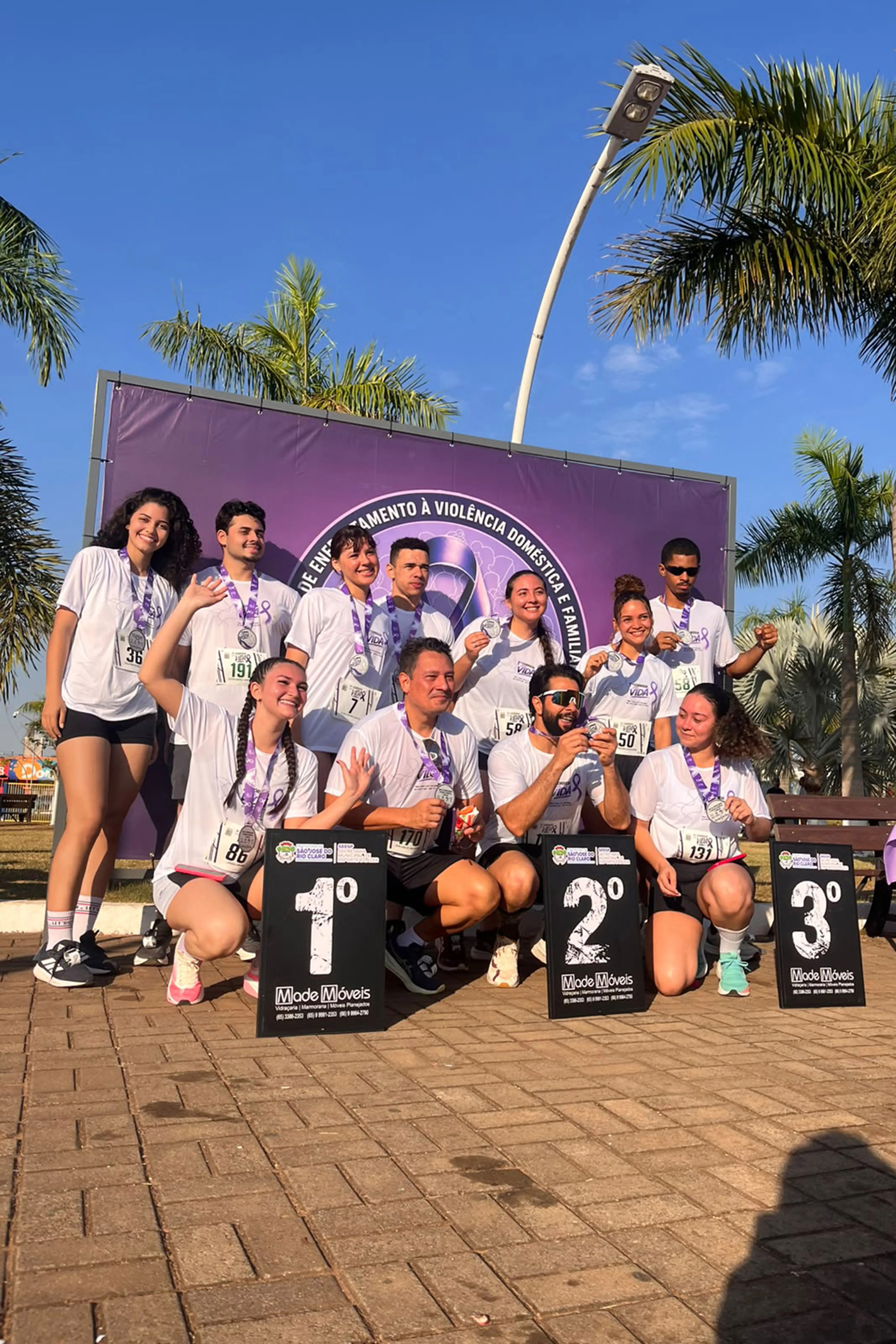 Grupo de participantes posa com medalhas ao pescoço diante de painel sobre enfrentamento à violência doméstica, ao lado de placas indicando as colocações do pódio.