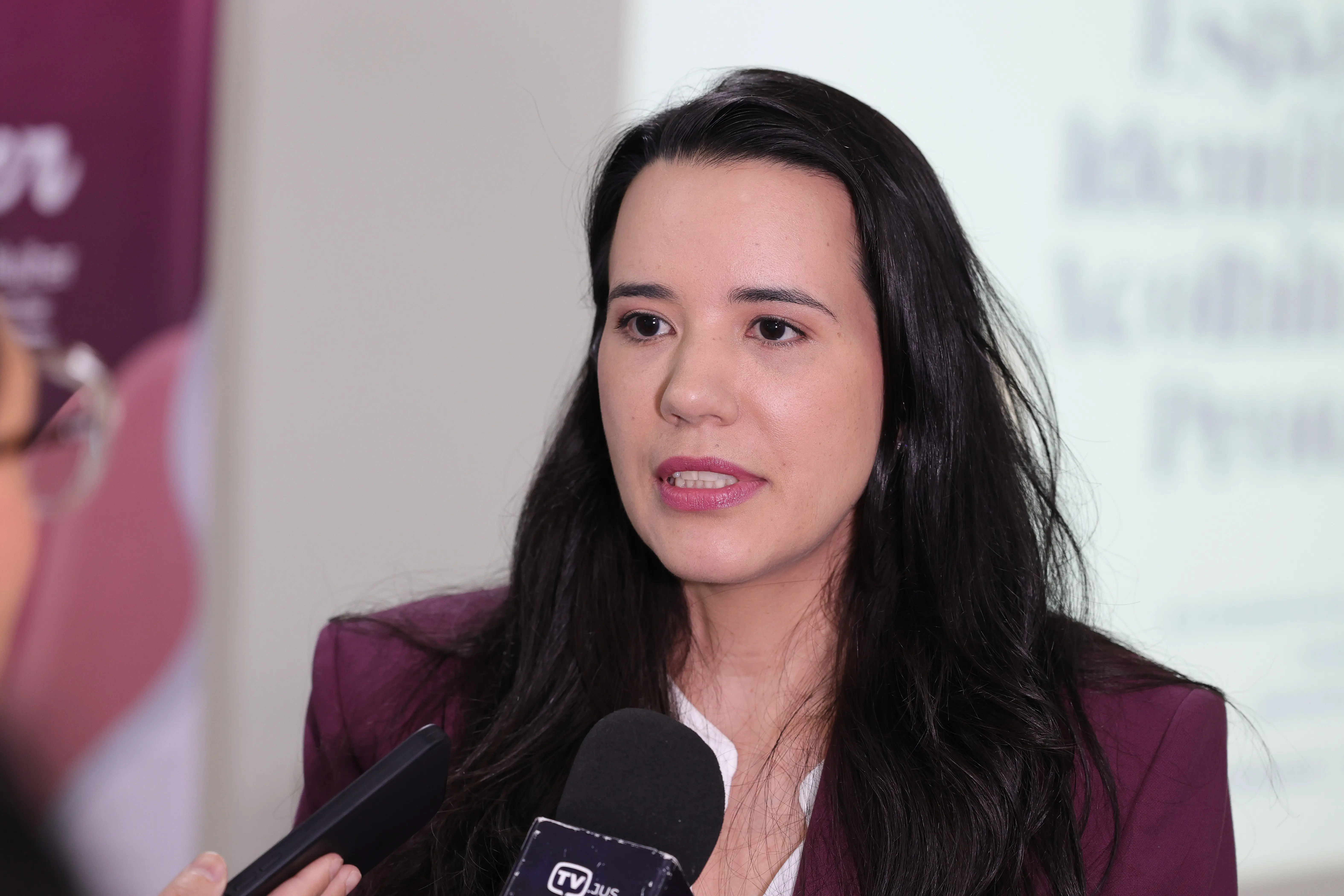 Mulher de cabelos escuros e blazer roxo fala ao microfone da TV Jus em uma entrevista. Ao fundo, um banner em tom de rosa e um telão com texto desfocado.