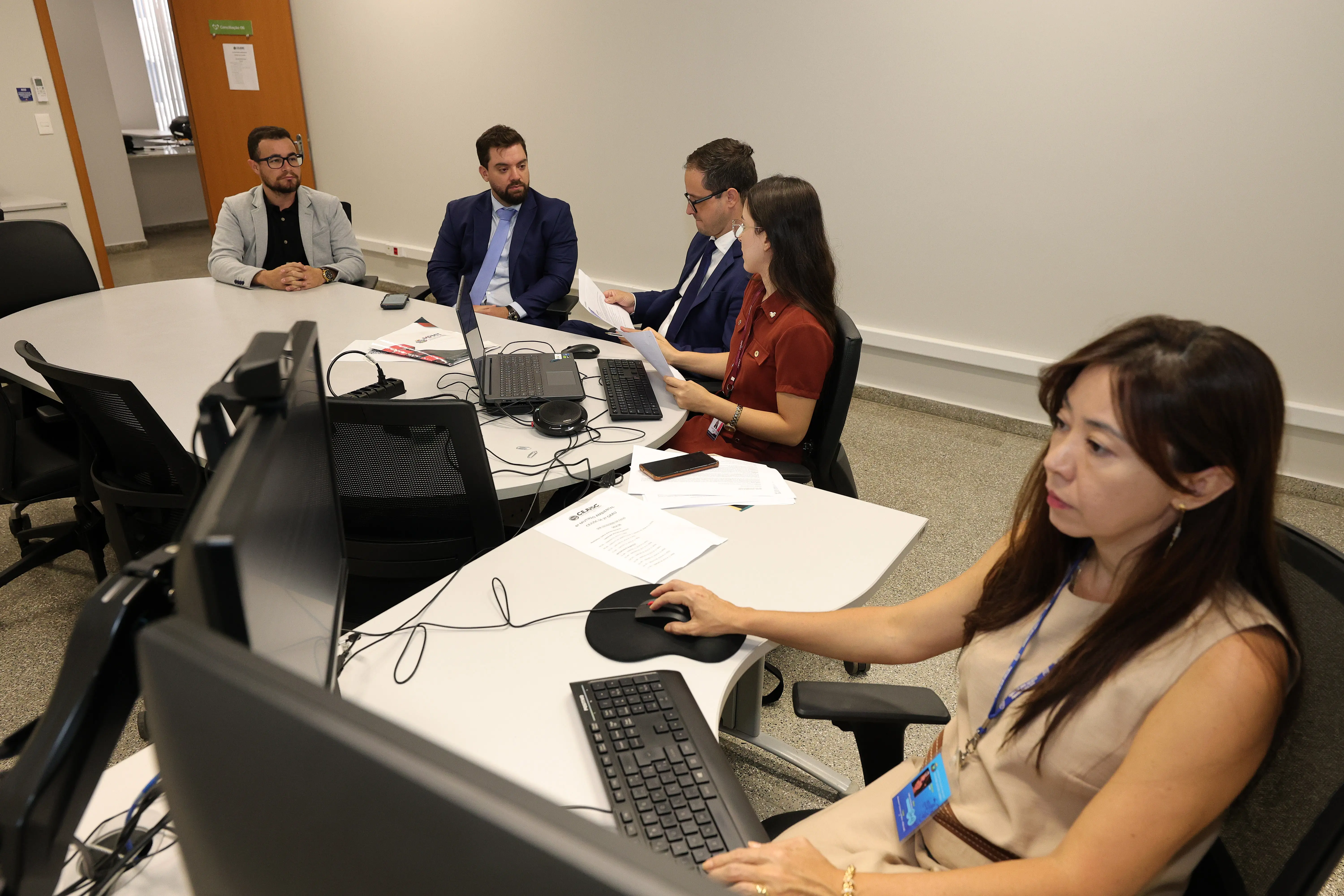 Vista em plano médio de uma sala de conferências onde cinco pessoas estão reunidas em torno de mesas brancas modulares. Três homens de terno e uma mulher de blusa vermelha conversam e olham para papéis. Outra mulher, em primeiro plano à direita, de blusa bege, digita no computador.