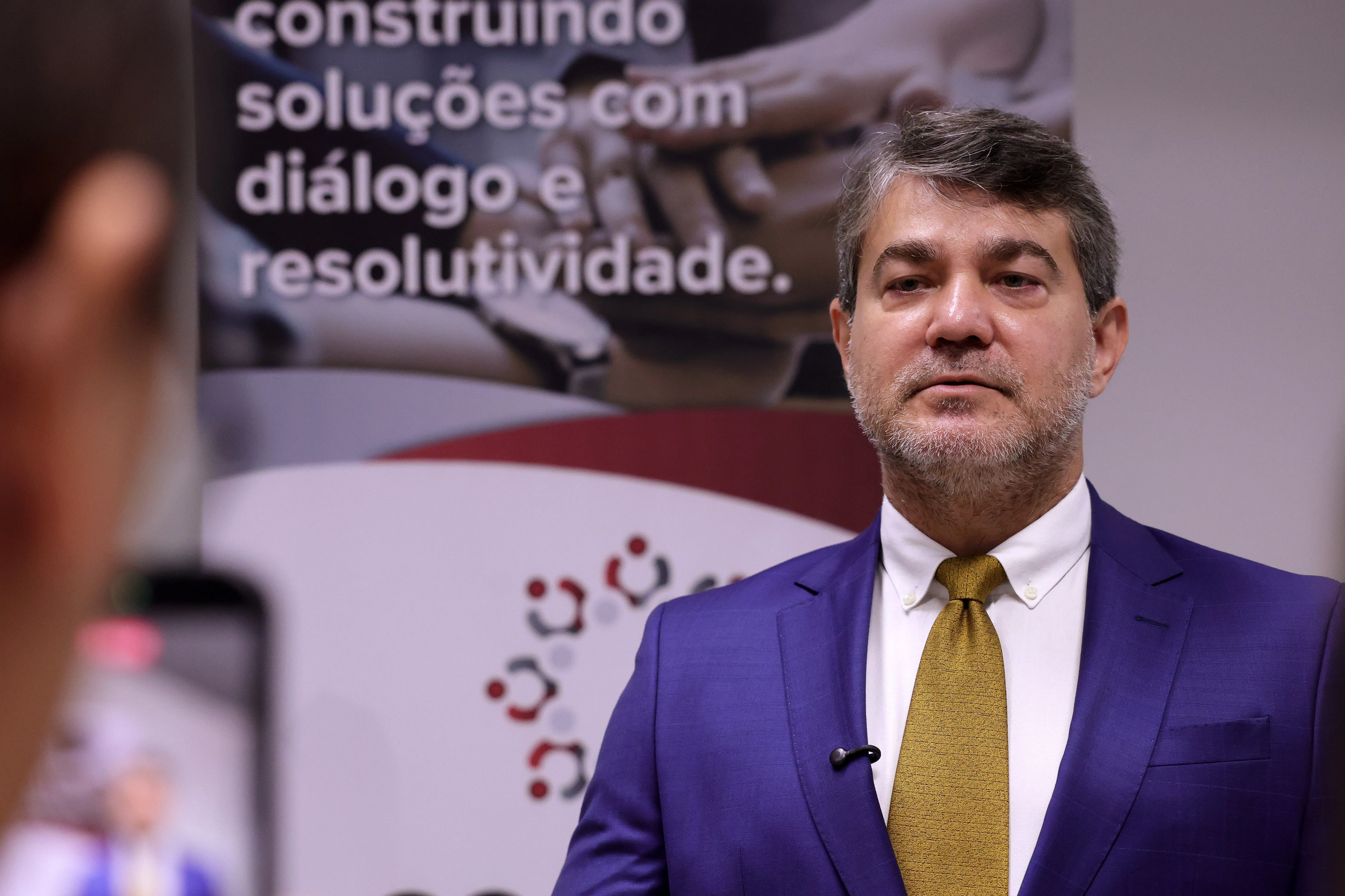 Retrato em plano fechado de um homem de meia-idade olhando para a direita. Ele tem cabelos grisalhos e barba por fazer e veste um terno azul royal com camisa branca e gravata amarela texturizada. Um microfone de lapela está preso na sua camisa. Ao fundo, um cartaz desfocado.