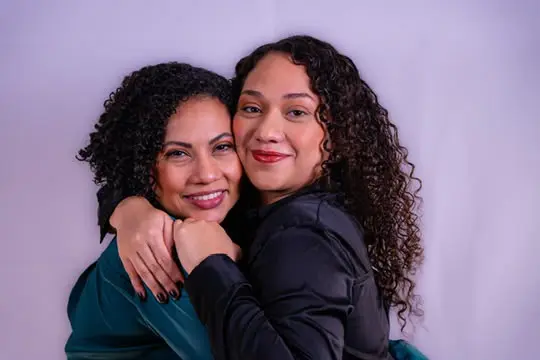 Duas mulheres de cabelos cacheados se abraçam e sorriem para a foto. Uma veste blusa verde-escura; a outra, blusa preta e batom vermelho.