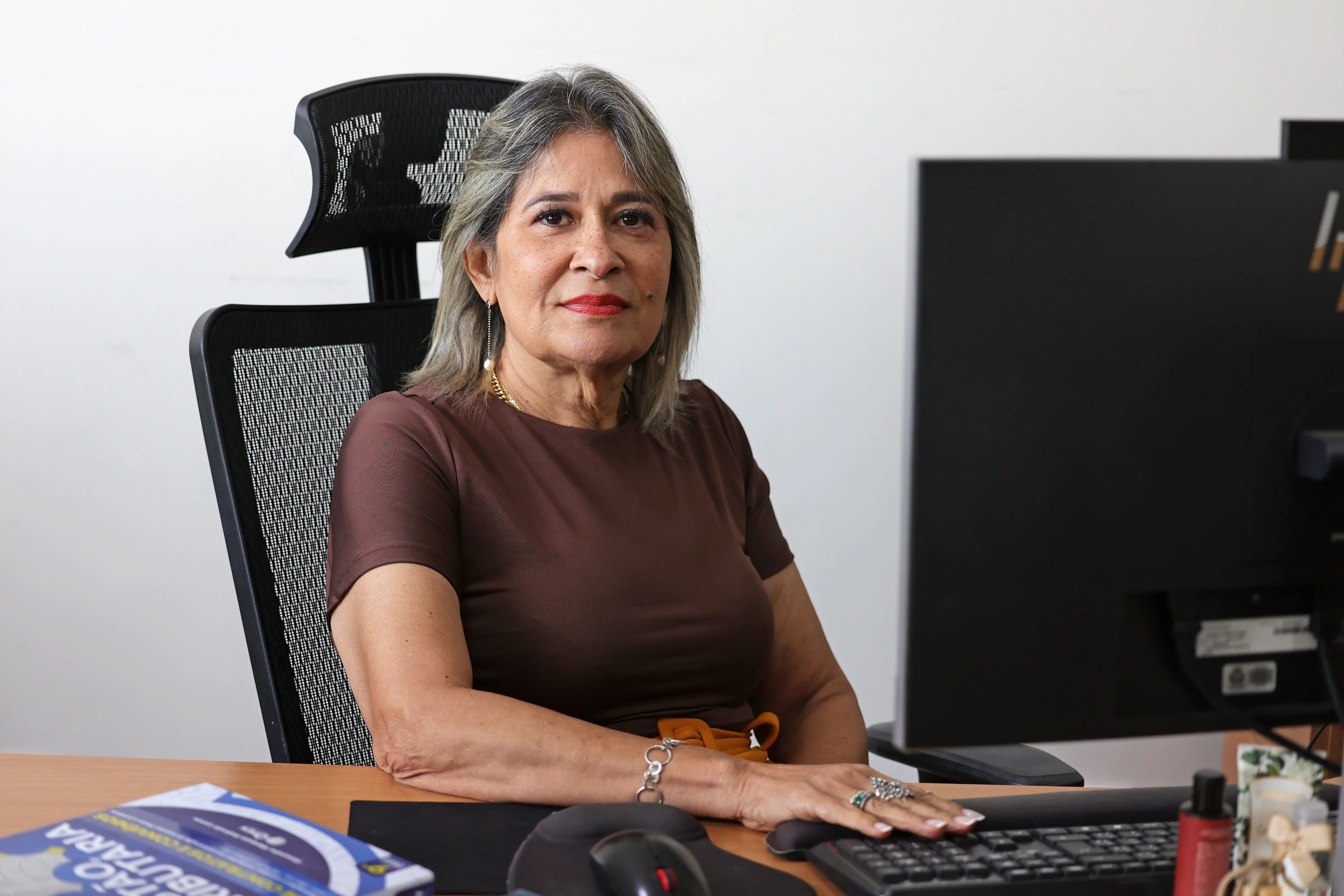Mulher de cabelos loiros e batom vermelho, blusa marrom, está sentada à mesa de trabalho, com a mão sobre o teclado, diante de um computador.