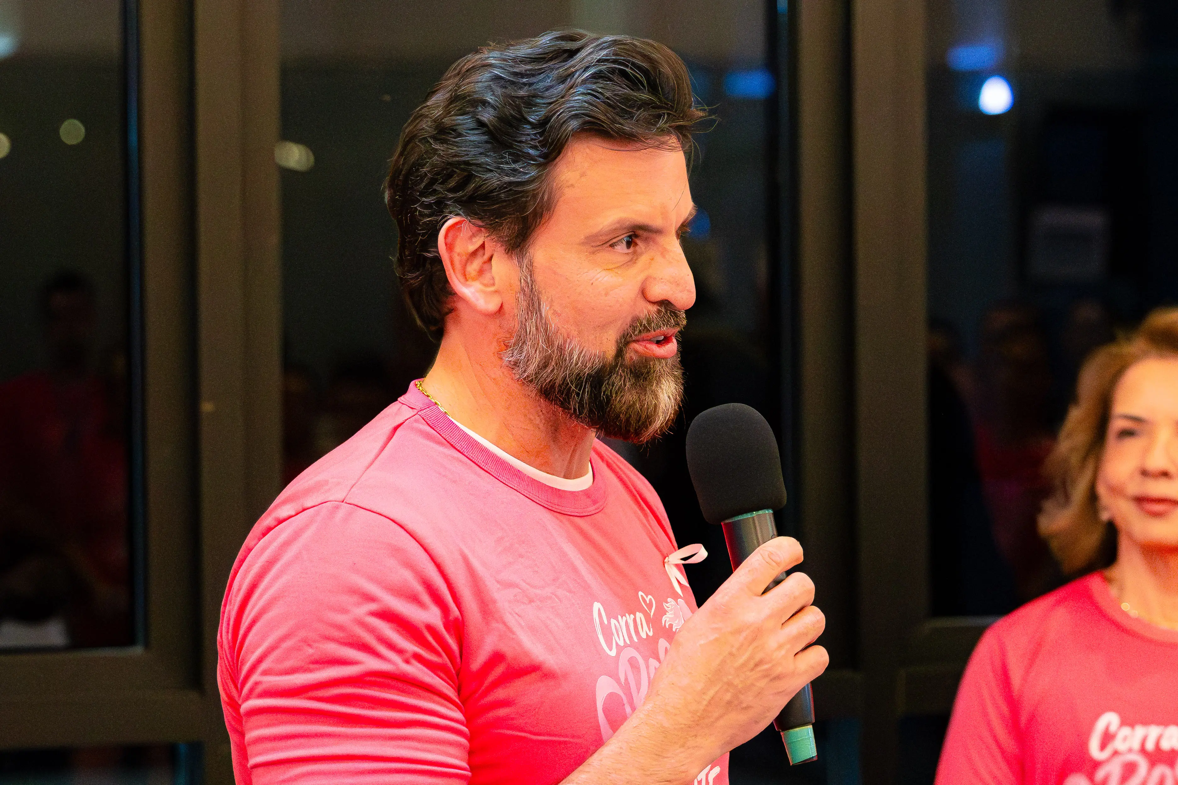 Homem barbudo de perfil fala ao microfone segurando-o com a mão, vestindo blusa rosa estampada. Parte de outra pessoa com blusa semelhante aparece ao lado direito.