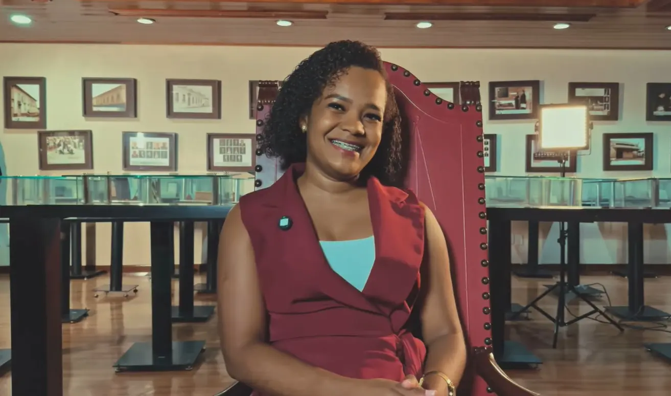 Foto horizontal que mostra a servidora Jéssica Silva sentada e sorrindo para a foto. Ela é uma mulher jovem, negra, de cabelo cacheado e castanho, usa blusa branca e colete vermelho. ela está no Espaço Memória do TJMT, um local com luz baixa e repleto de quadros e documentos antigos expostos.