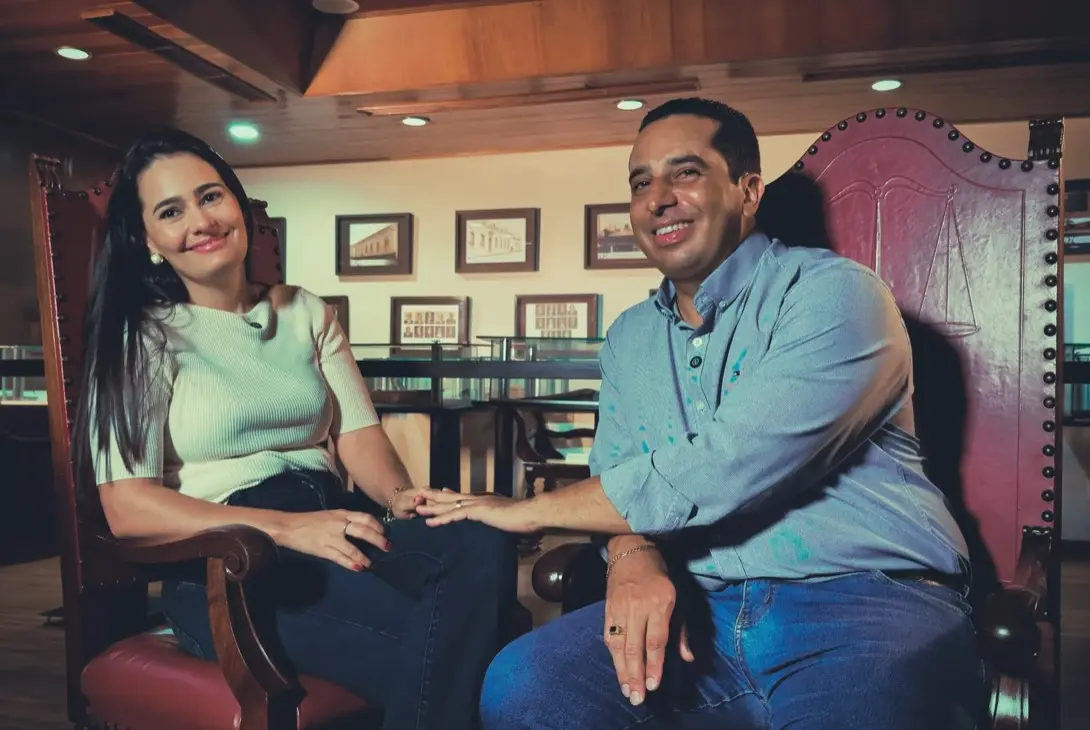 Foto horizontal que mostra o casal de servidores Valdete e Sérgio Rangel sentados em cadeiras, sorrindo. Ele coloca a mão esquerda sobre a mão dela, que está repousada sobre a coxa. Eles estão no Espaço Memória do TJMT, um local com luz baixa, repleto de quadros e documentos antigos expostos.