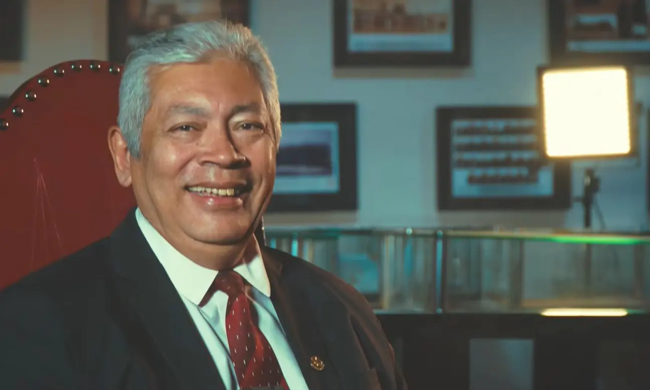 Foto horizontal que mostra o servidor Dito Gonçalo sentado, sorrindo para a foto. Ele é um senhor negro, de cabelos lisos e grisalhos, usa camisa branca, gravata vermelha e terno preto. Ele está no Espaço Memória do TJMT, um local de luz baixa, com quadros e documentos antigos expostos.