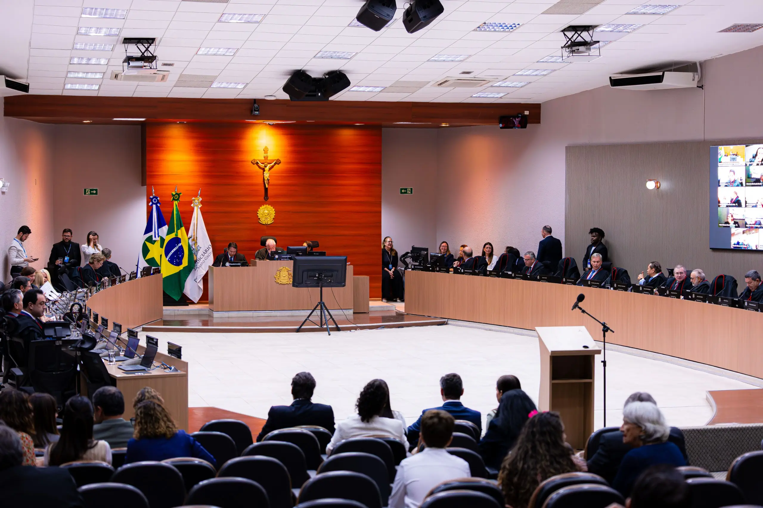 Plenário judicial amplo com mesa em semicírculo onde autoridades se sentam. Ao fundo, parede de madeira com crucifixo e bandeiras. Em primeiro plano, plateia assiste à sessão.
