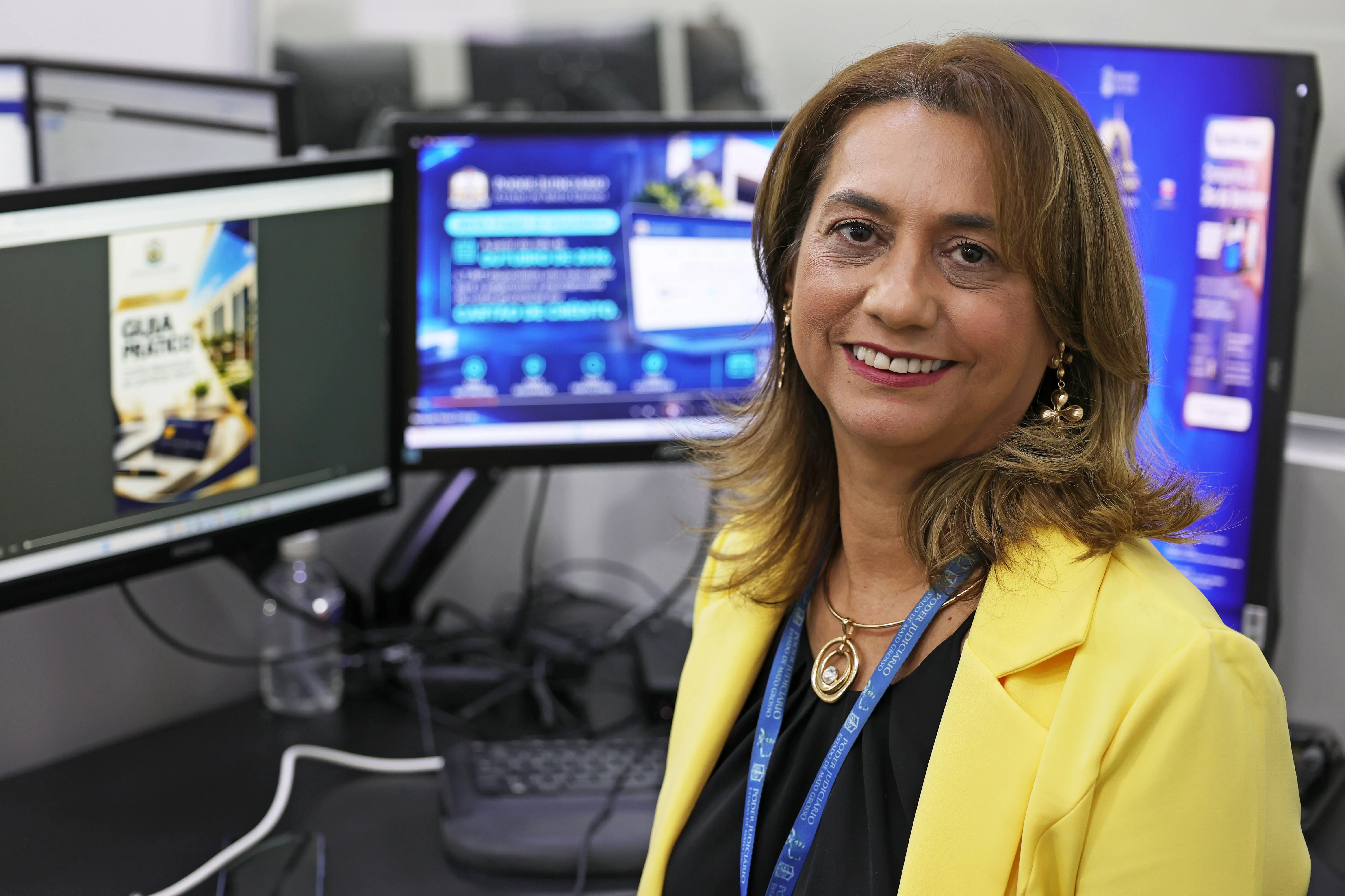 Mulher de cabelos castanhos, sorridente, veste blazer amarelo e cordão azul do Poder Judiciário. Atrás dela, monitores exibem material de divulgação do TJMT.
