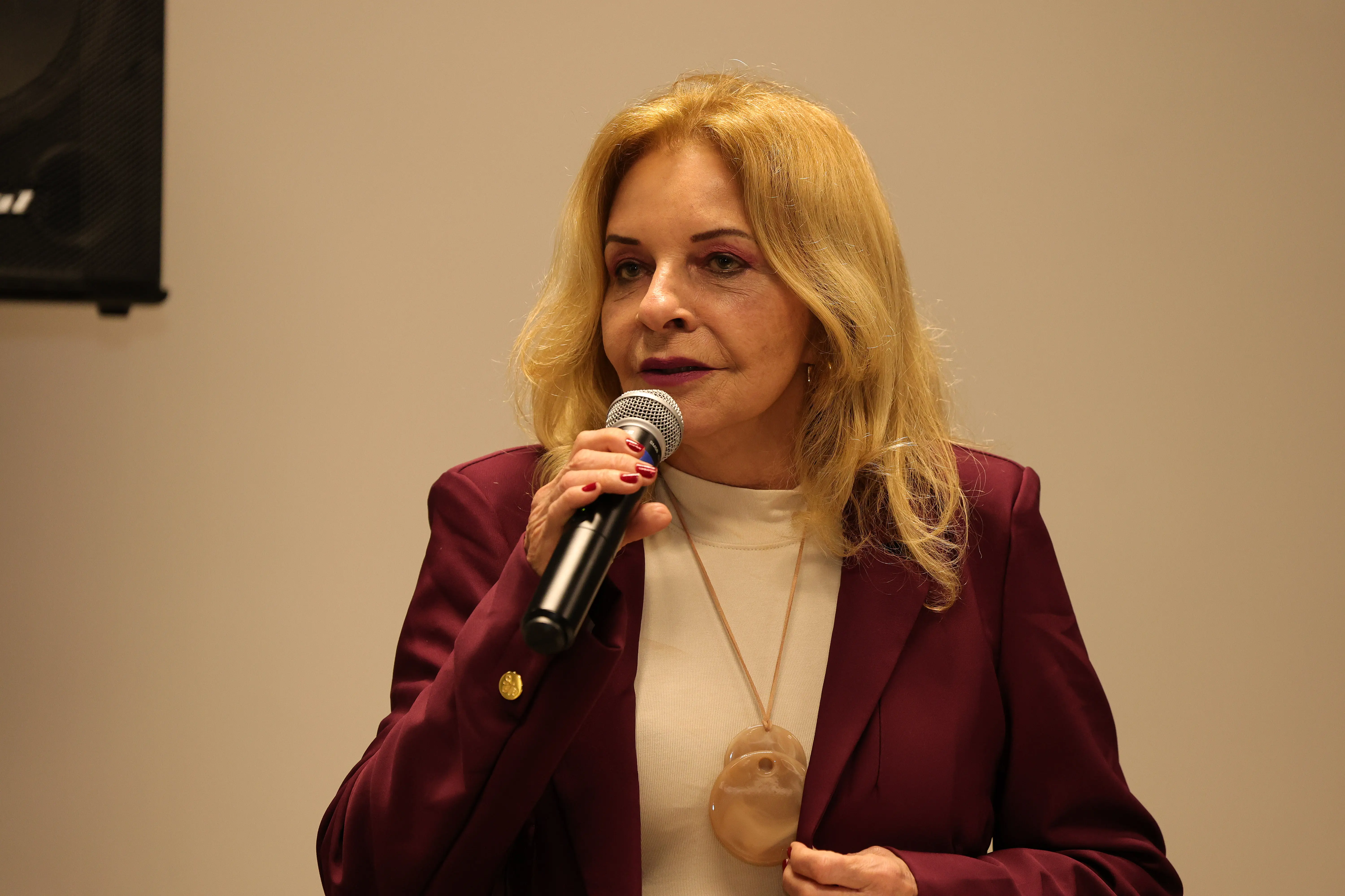 Fotografia de meio-corpo de uma mulher loira de cabelos ondulados falando ao microfone com o olhar voltado para a frente. Ela usa um blazer vinho sobre uma blusa branca de gola alta e um colar com pingente redondo grande e claro. Unhas pintadas de vermelho.