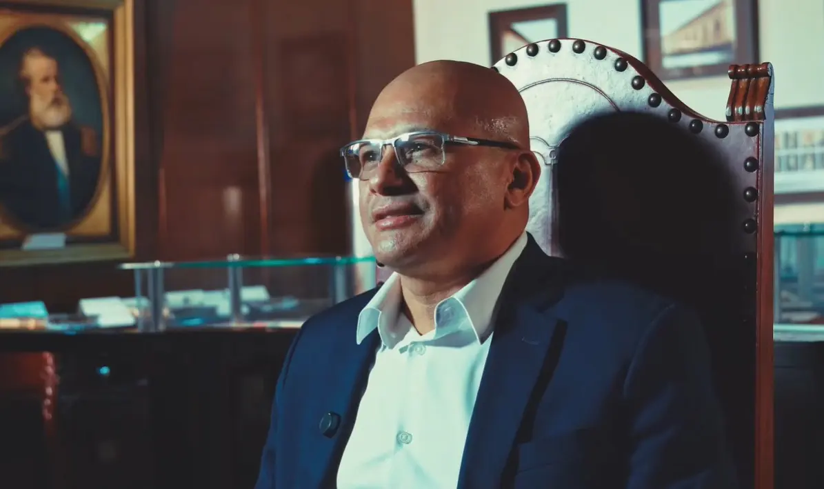 Foto horizontal que mostra o servidor Amarildo Gonçalo sentado em uma cadeira de madeira. Ele é um homem negro, careca, usa camisa branca e blazer azul marinho. Ele está no Espaço Memória do TJMT, um local com luz baixa, quadros, móveis antigos e documentos expostos.