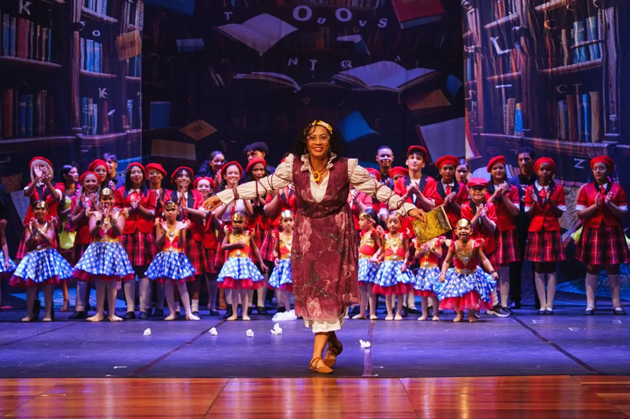 Mulher de óculos, colete vinho e tiara dourada abre os braços em palco decorado com estantes de livros. Ao fundo, crianças de boina vermelha e saias xadrez ou azuis aplaudem.