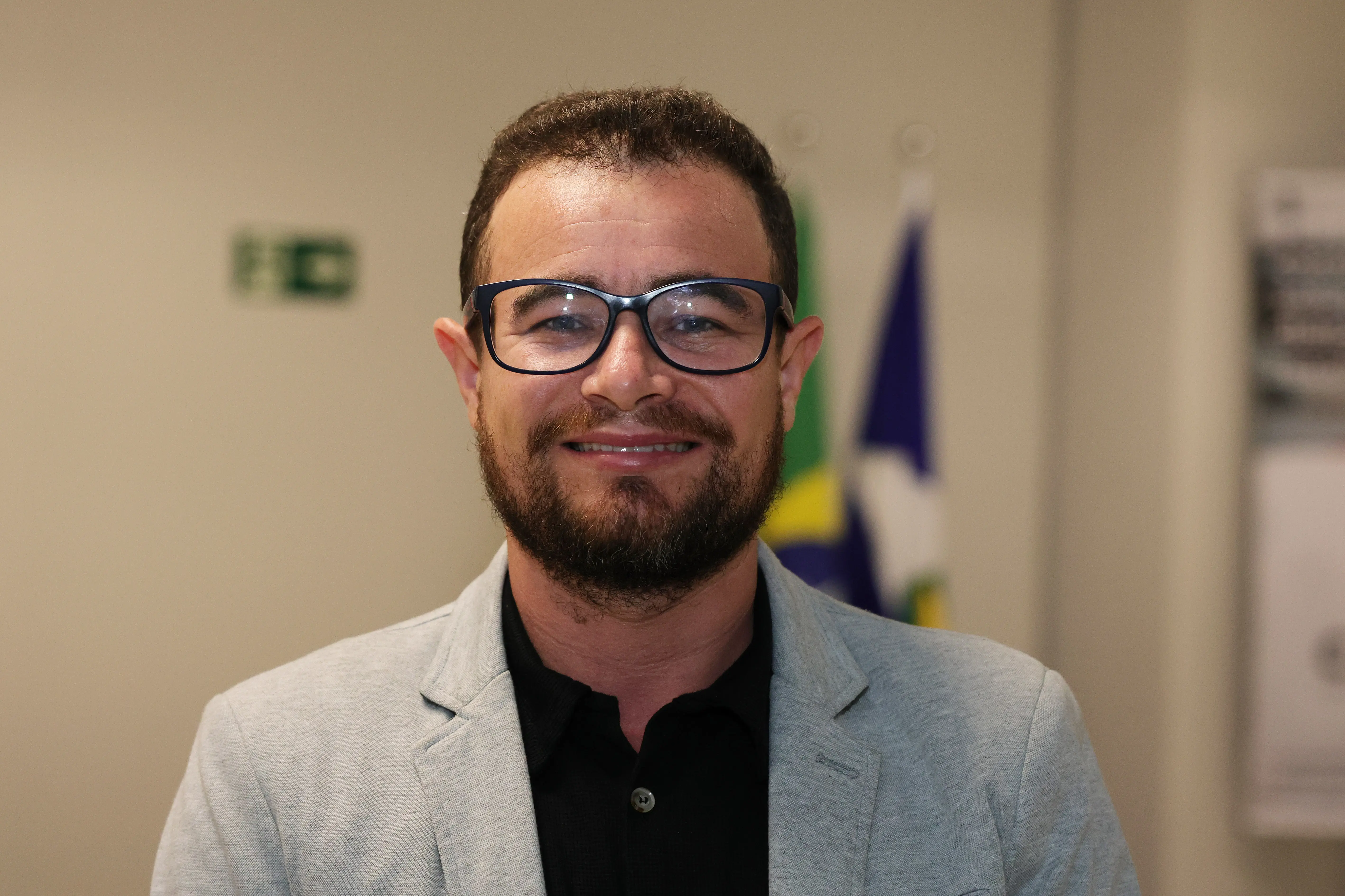 Fotografia de meio-corpo de um homem com óculos pretos, barba cheia e cabelos escuros, sorrindo e olhando para a câmera. Ele veste um blazer cinza claro sobre uma camisa social preta. Ao fundo, desfocados, há mastros de bandeiras e uma sinalização de saída de emergência verde.