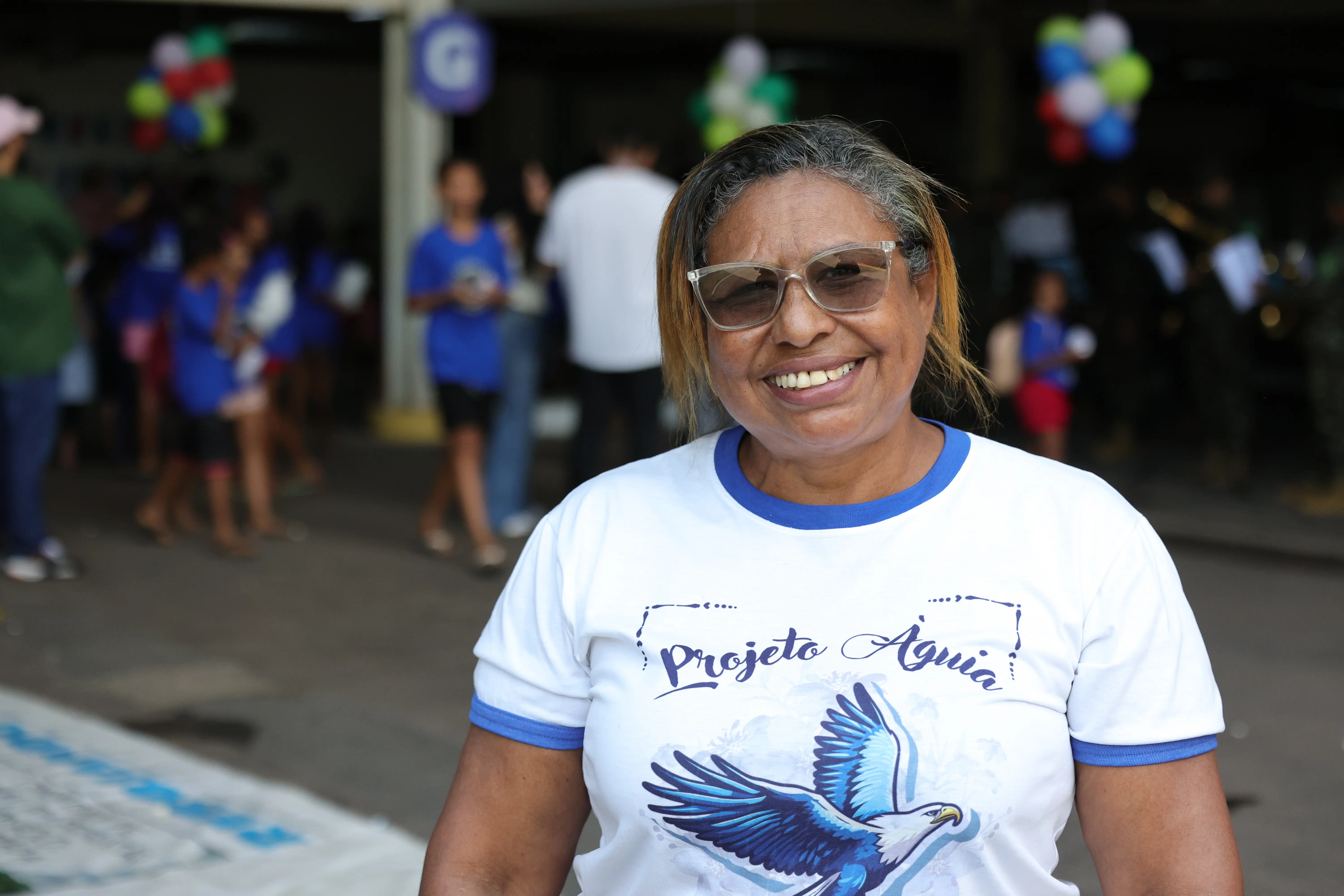 Mulher sorridente de óculos usa camiseta branca do "Projeto Águia" estampada com uma ave azul. Ao fundo desfocado, pessoas transitam sob balões coloridos suspensos.