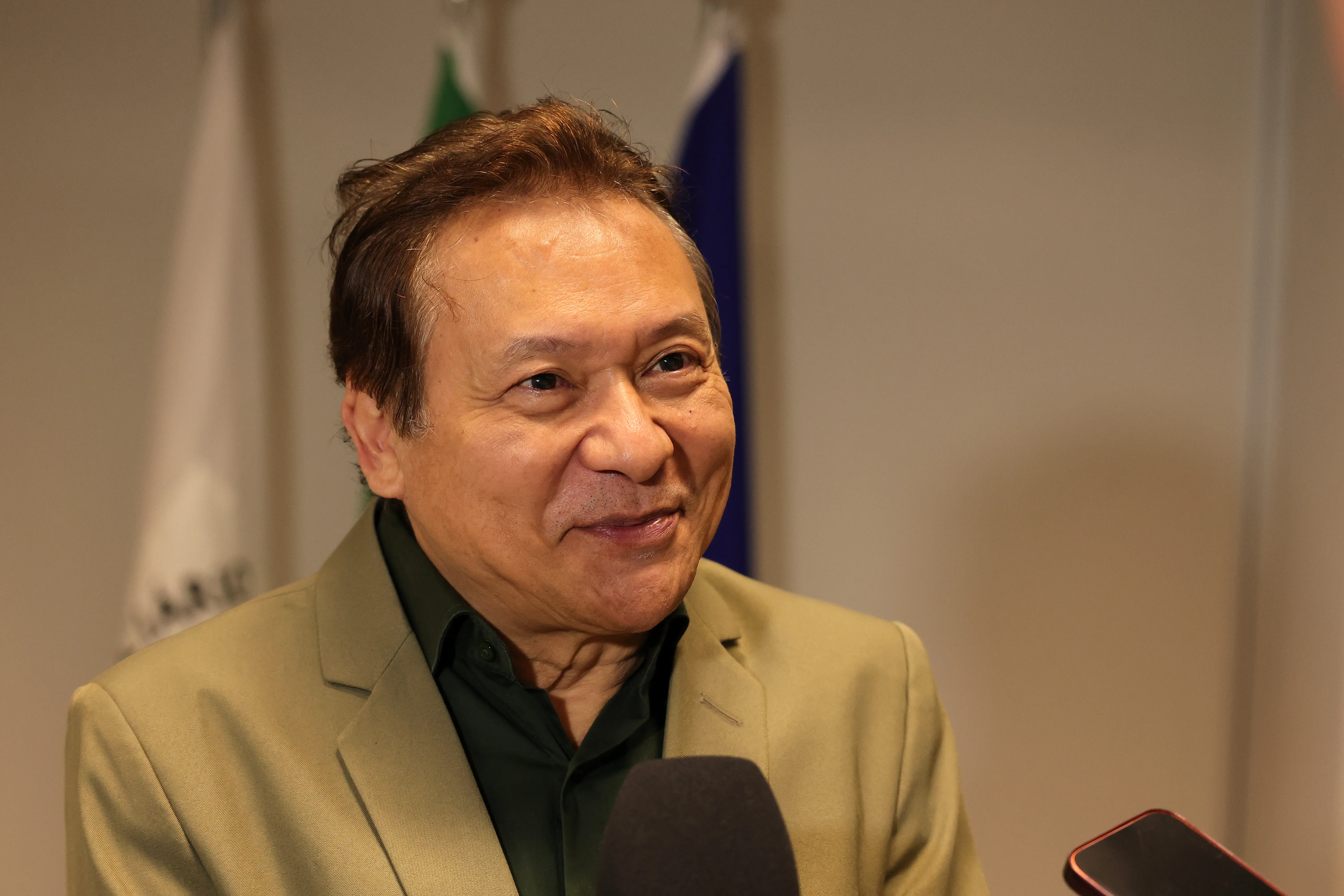 Fotografia em plano fechado do rosto de um homem idoso sorrindo e olhando ligeiramente para a direita. Ele tem pele morena, cabelos escuros e veste um blazer verde oliva sobre uma camisa verde escura. Um microfone está próximo a ele na parte inferior.
