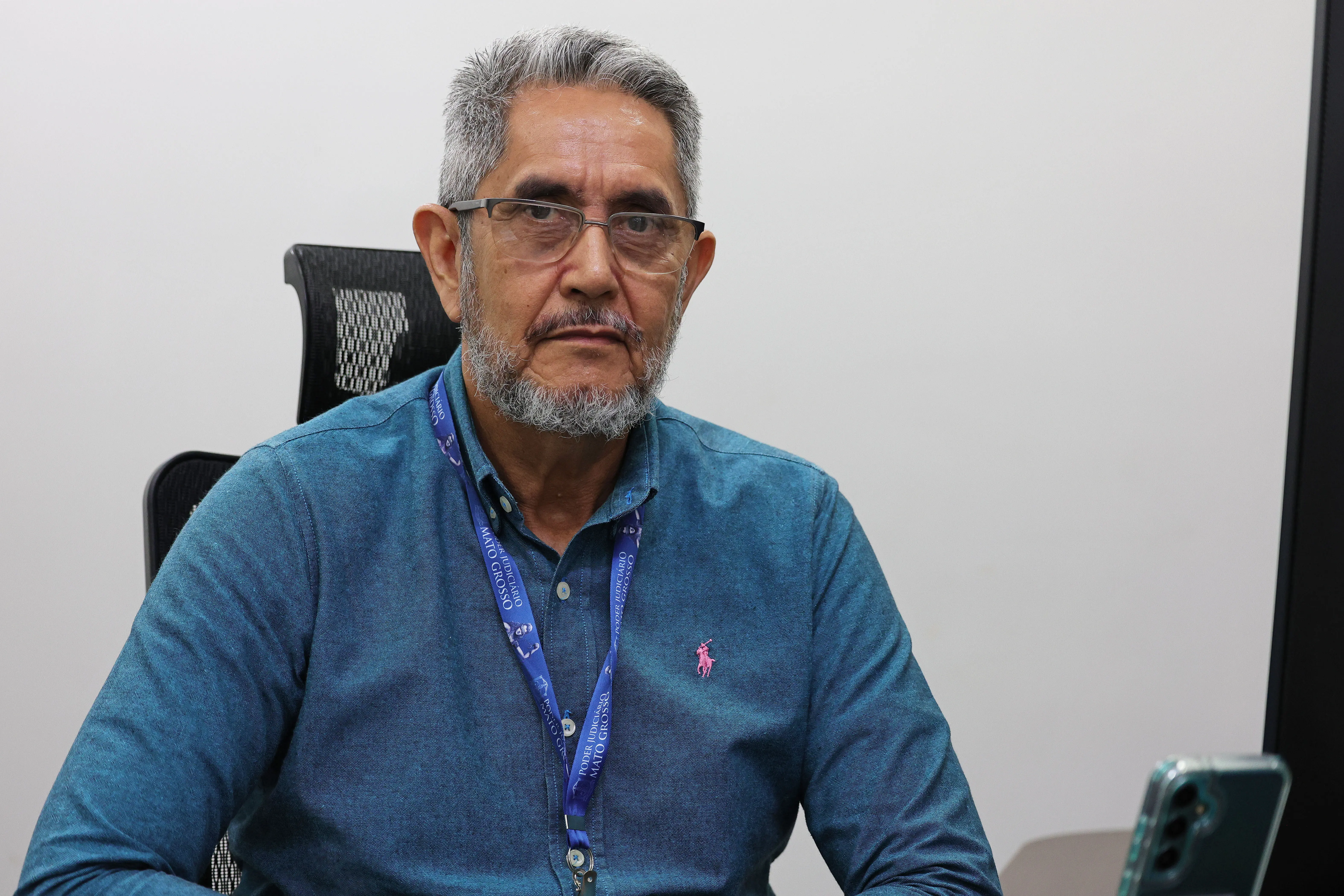 Homem de cabelos e barba grisalhos, óculos e camisa azul, usa cordão do Poder Judiciário de Mato Grosso e está sentado em cadeira de escritório. 