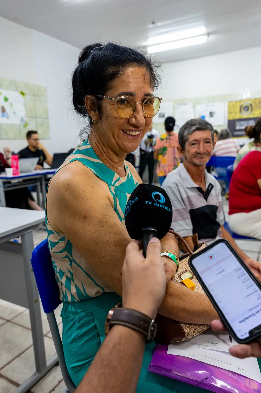 Mulher parda sorridente, de óculos e cabelos presos, usa blusa verde e bege. Ela é entrevistada com microfone da TV Justiça e celular gravando. Ao fundo, um homem e ambiente de escritório.